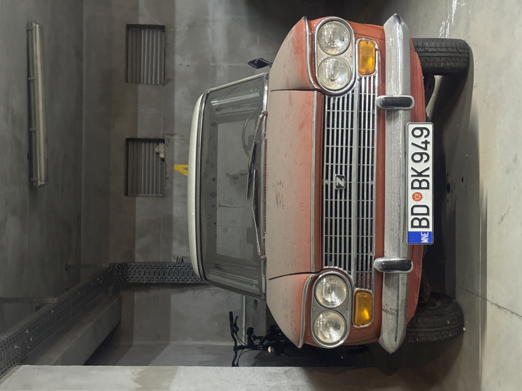 An eye-level, full shot captures a dusty, faded red and white vintage car parked indoors in what appears to be a concrete garage in Budva, Montenegro. The car, positioned centrally, faces forward, its headlights on. The front of the car features a chrome grille with a "Z" emblem, two large circular headlights, and amber turn signals. A license plate reading "MNE BD BK 949" is mounted on the chrome bumper. To the left of the car, a dark bicycle is partially visible. The garage environment is characterized by rough concrete walls, a concrete ceiling with exposed conduits and a fluorescent light fixture, and two louvered windows. The floor is also concrete, with some debris or water stains present. The overall impression is one of a dimly lit, utilitarian space housing an old vehicle.