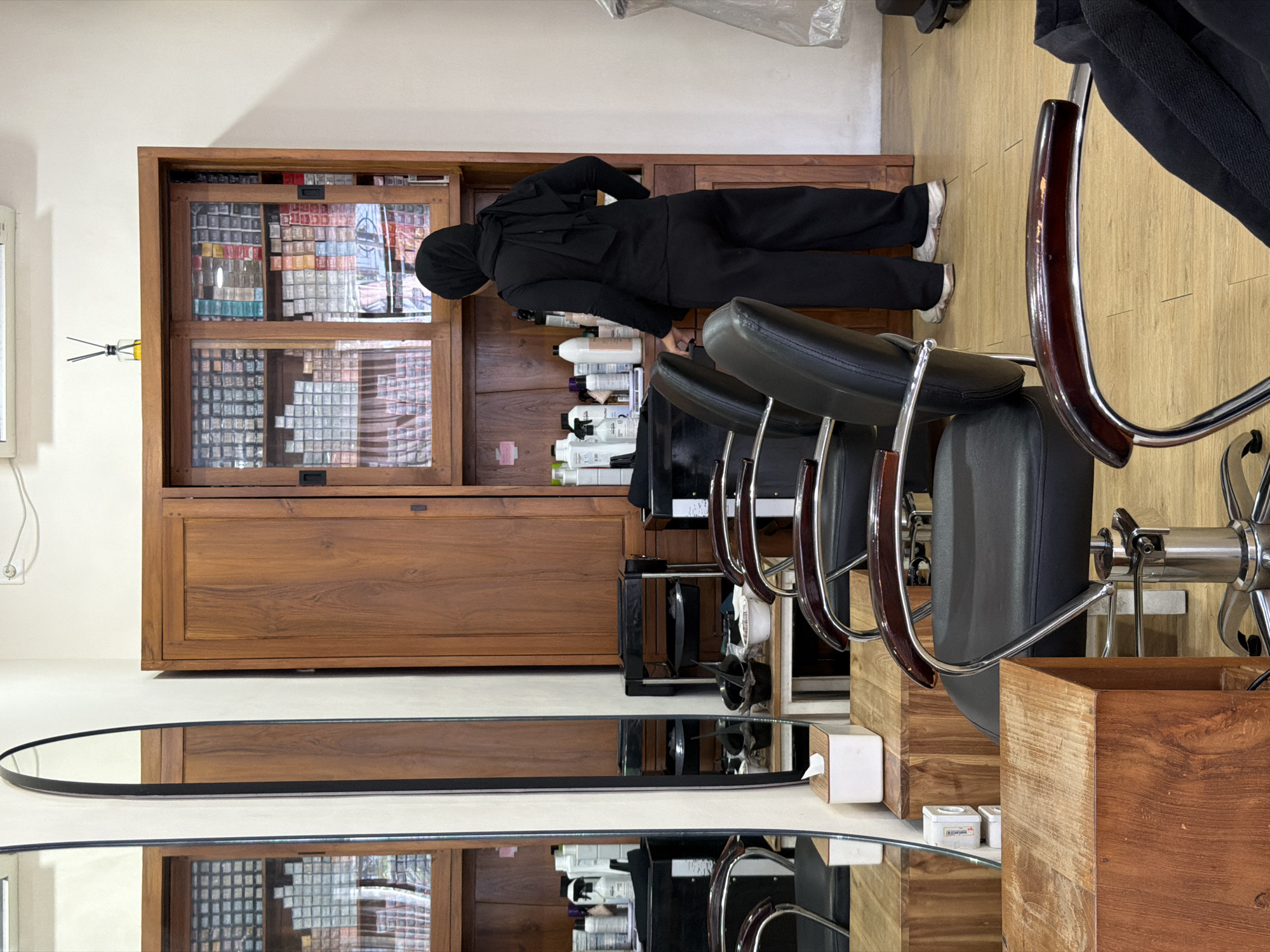 A person wearing a black hijab and dark clothing is reaching into a wooden cabinet containing multiple small boxes in what appears to be a salon setting in North Kuta, Indonesia. The cabinet holds numerous boxes, possibly hair dye or other salon supplies. Rows of salon chairs with dark upholstery and chrome bases are visible, along with an oval mirror placed near the base of the cabinet. The floor is light-colored with a wood grain pattern. Bottles of various products are arranged on the shelf within reach of the person. The scene is indoors and suggests a professional salon environment.