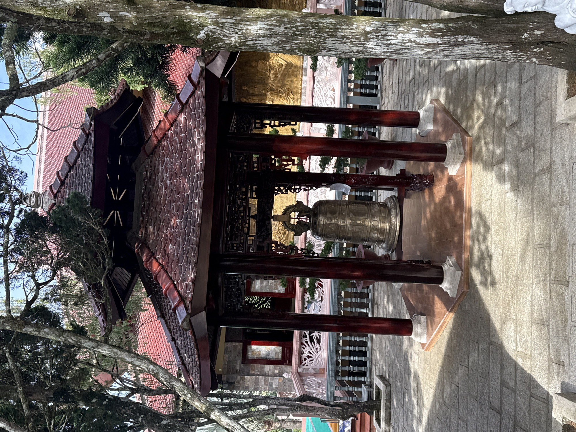 This is a full, eye-level, outdoor, daytime, medium shot of a large bell housed in a hexagonal wooden pavilion in Da Lat, Vietnam. The pavilion has a dark red, multi-tiered, tiled roof with upturned eaves, and is supported by four dark red wooden pillars with white bases. The bell itself is large, made of brass, and sits on a wooden platform within the pavilion. The ground is paved with light gray stone tiles. In the background, a building with a red tiled roof and stone walls is visible, with a decorative railing in front of it. Trees with rough bark and green foliage surround the scene, with some branches extending into the frame. The lighting suggests it is a sunny day, with dappled shadows cast on the ground.