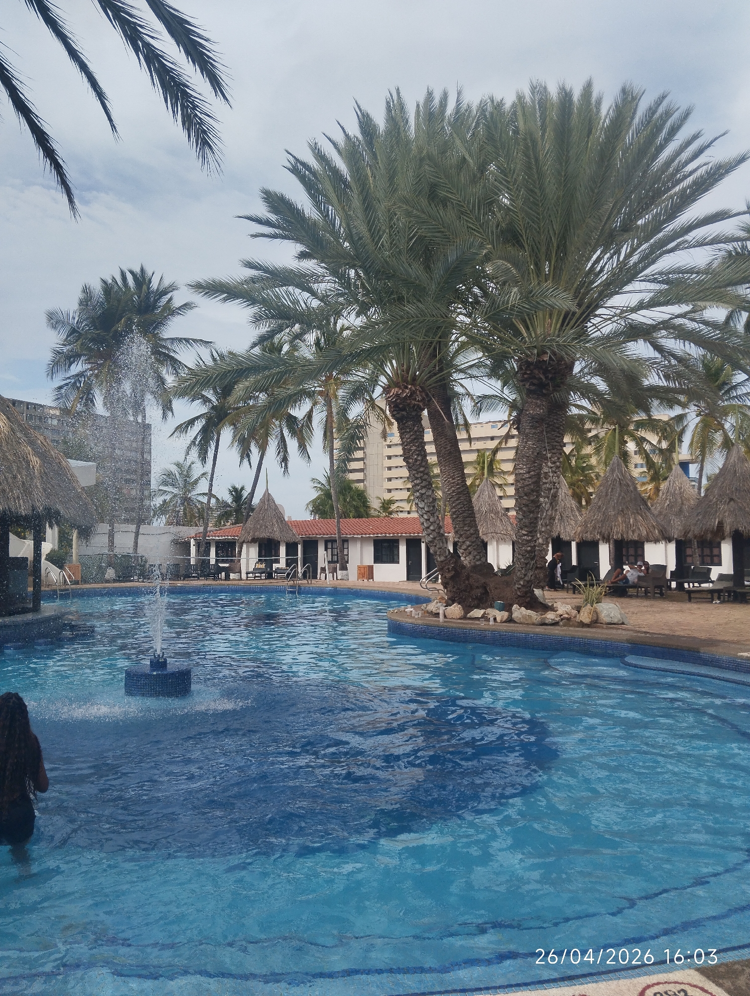 The image depicts a swimming pool area with palm trees and thatched-roof structures. The pool features a fountain in the foreground and a person standing in the water. In the background, there are buildings and additional palm trees. Several people are sitting under the thatched-roof structures. The location is Unknown, Unknown. A date stamp of 26/04/2026 16:03 is visible in the bottom right corner.