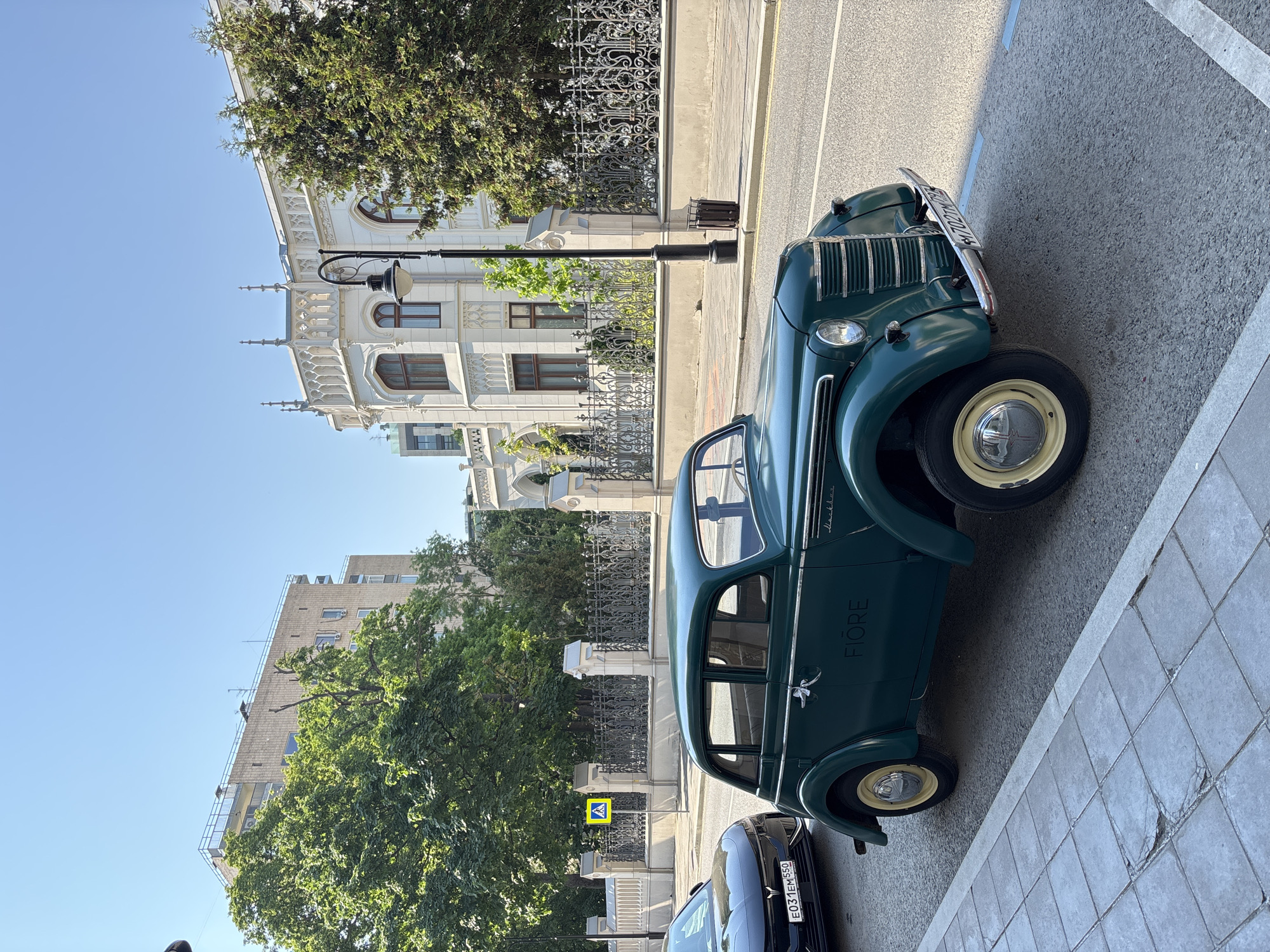An eye-level, outdoor shot shows a green vintage car parked on a city street in Moscow, Russia. The car is facing the right side of the frame. A modern black car is partially visible behind it. A pedestrian crossing sign stands near the vintage car. Behind the car is a wrought-iron fence. Buildings are visible behind the fence. The sky is clear blue. The lighting appears to be from bright sunlight.