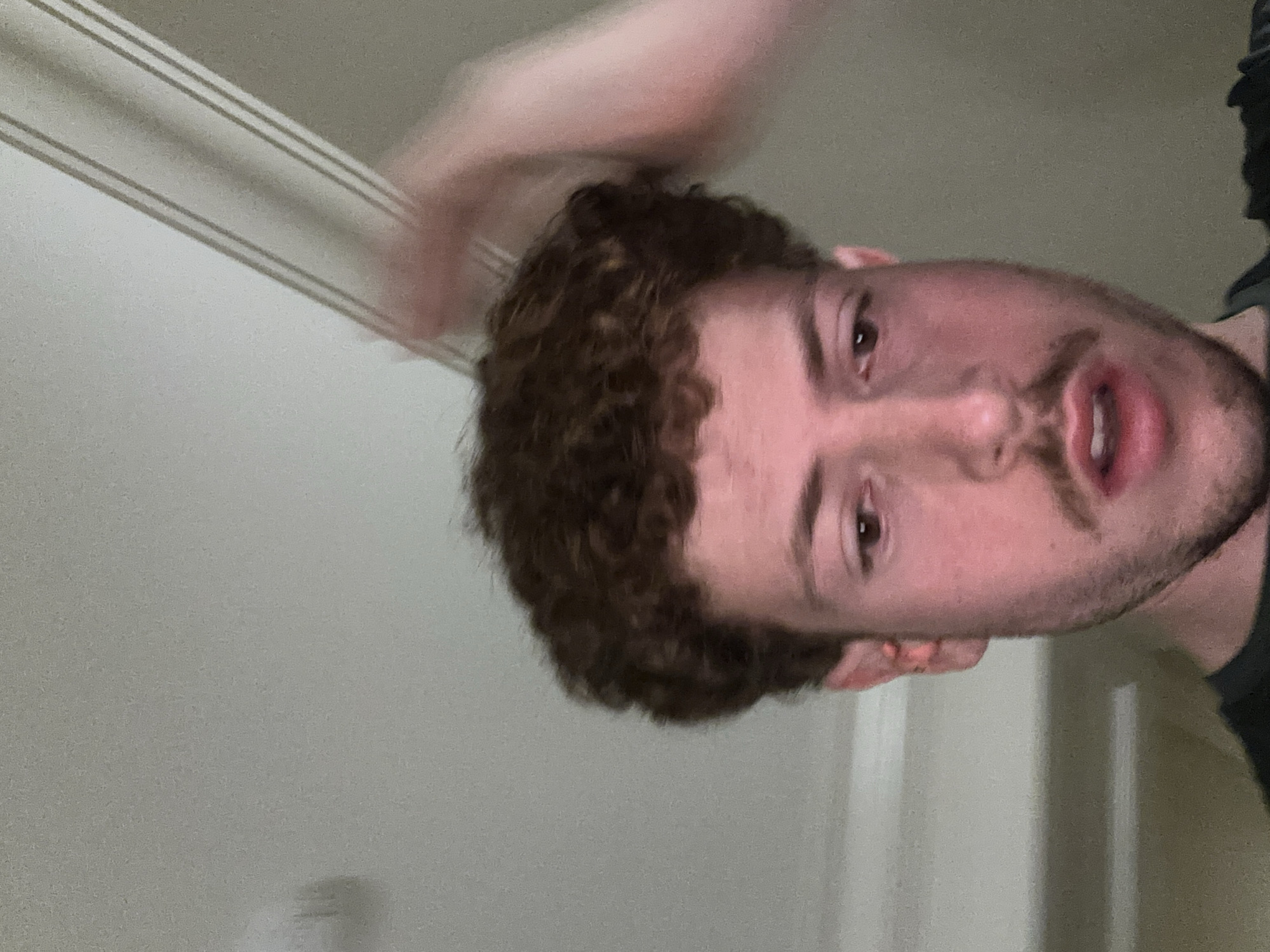 A medium close-up shot depicts a Caucasian male with brown curly hair, brown eyes, and a trimmed mustache. He is positioned towards the right side of the frame, with his face angled slightly upwards. His mouth is partially open. A blurred, indeterminate pale form is present in the upper portion of the image, above the subject's head. The background consists of a light-colored wall. The specific location in Aventura, United States of America cannot be determined from the given image.