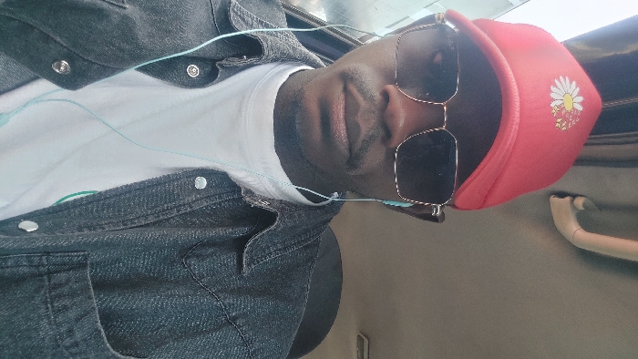 A male individual is depicted in a close-up portrait, seated inside what appears to be a vehicle in the city of Hoss, Nigeria. He wears a red baseball cap adorned with a white daisy design, dark rectangular sunglasses with gold-colored frames, and light blue wired in-ear headphones. His attire consists of a white t-shirt layered under an unbuttoned dark denim-style jacket with silver snap buttons. A mustache is visible above his upper lip. The interior background shows a light-colored headliner, a beige grab handle, and ambient light entering from a window on the left.