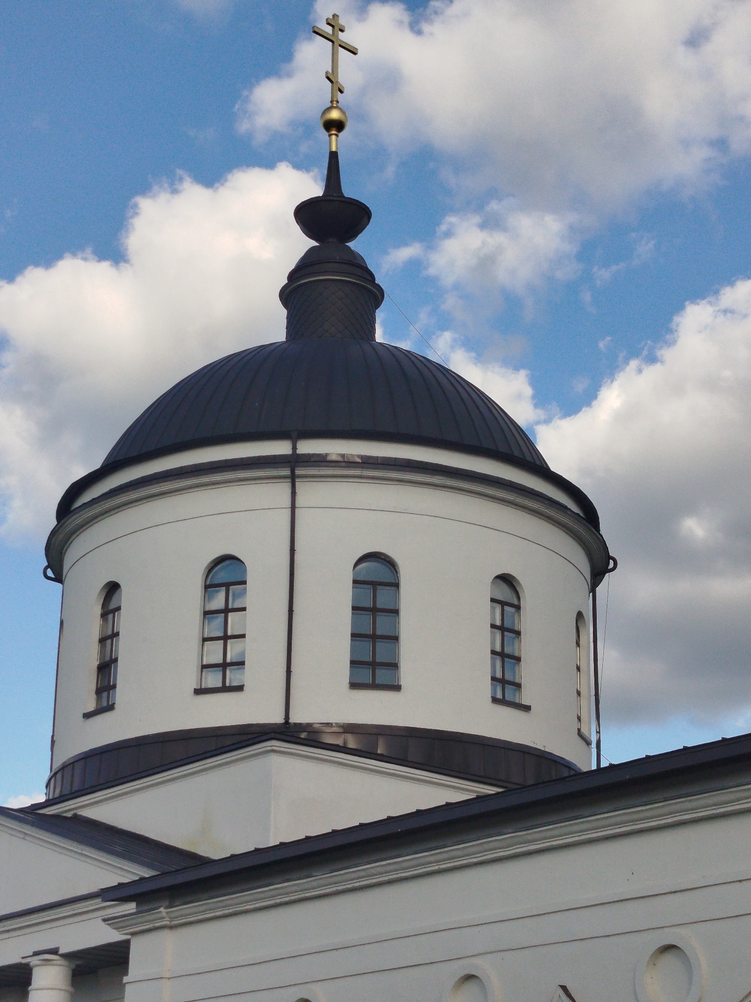 The image shows a close-up of the dome of a Russian Orthodox church in Боровково, Russia. The dome is white with a dark gray or black roof and a gold cross on top. There are several arched windows on the dome, each with a multi-pane window. The sky behind the dome is blue with white clouds. The architecture is classical with clean lines.