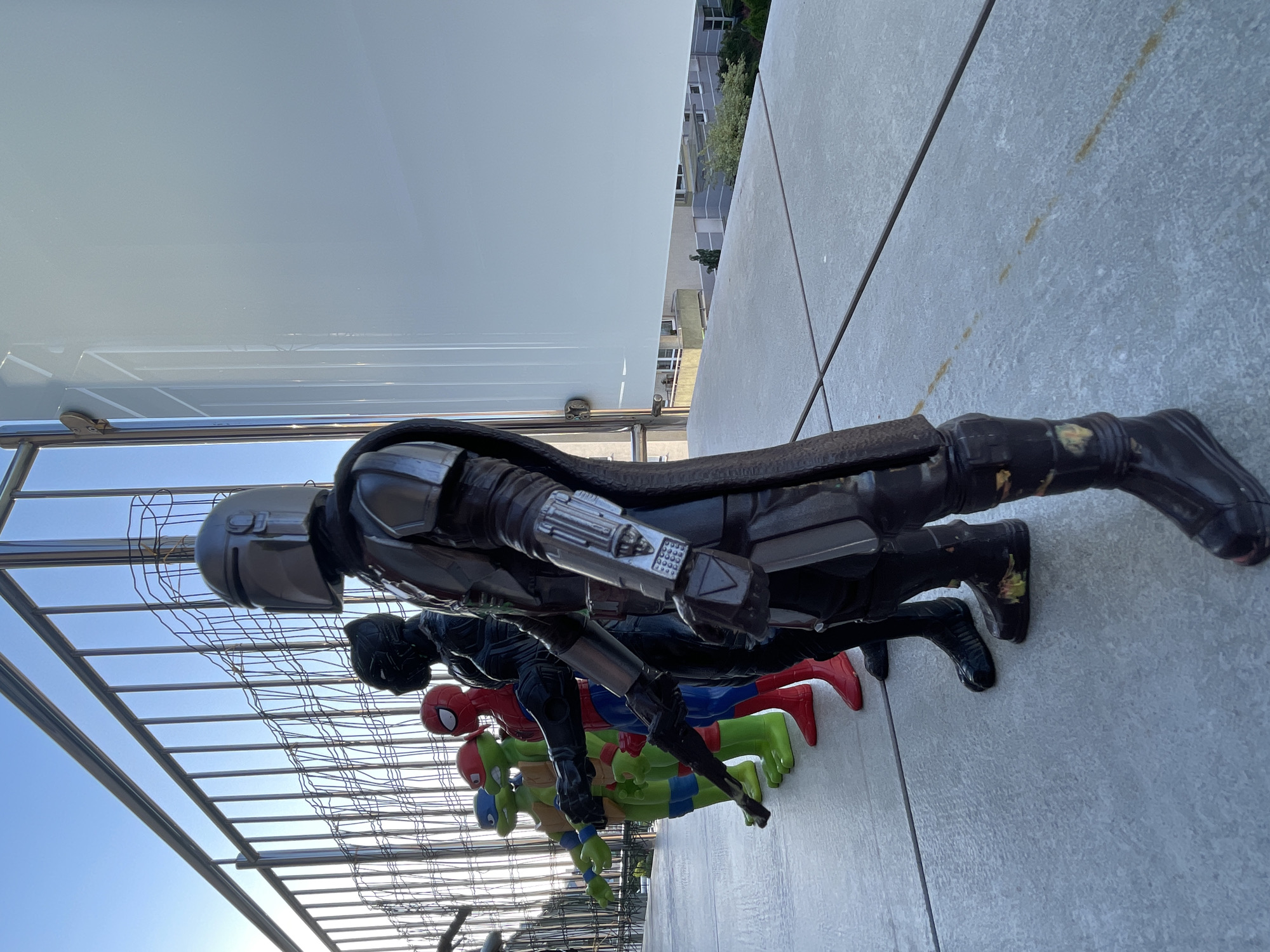 A line of action figures stands on a concrete patio against a bright blue sky. From foreground to background, the figures are: The Mandalorian, Black Panther, Spider-Man, Raphael of the Teenage Mutant Ninja Turtles, and Michelangelo of the Teenage Mutant Ninja Turtles. The figures are positioned facing right, their arms extended forward. The Mandalorian figure is in the front and right of the image, with its metallic helmet and dark armor dominating the foreground. Behind it are the other figures, each in their distinct superhero costumes. The figures are standing in a row, their feet on the grey, tiled concrete. A silver railing runs horizontally along the top of the image, with thin metal vertical bars creating a fence-like structure. To the right of the figures, a large, smooth, white panel extends upwards, reflecting some of the light. In the background, beyond the railing and fence, residential buildings are visible, suggesting an urban or suburban setting. The overall lighting indicates it is daytime, likely late afternoon or early morning due to the angle of the sun casting long shadows. The concrete patio appears to have some faint reddish-brown marks on its surface.
