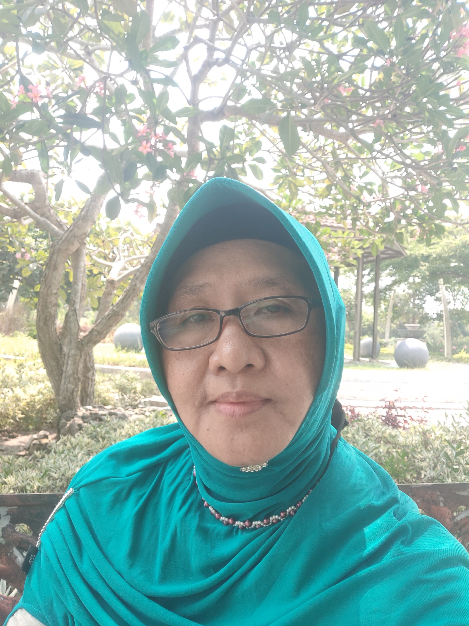 A woman wearing a teal hijab and glasses is captured in a selfie. She has medium-toned skin and is looking directly at the camera. The woman is wearing a beaded necklace. In the background, there are trees with pink flowers and foliage, suggesting an outdoor setting with lush greenery. Some park-like structures and round, grey objects are also visible in the distance, possibly indicating a park or garden area. The scene appears to be illuminated by natural sunlight. The provided context suggests the location is Pagu, Indonesia.