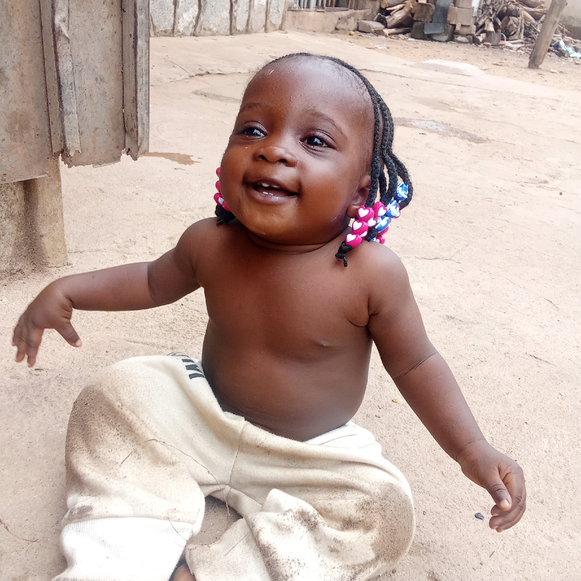 A young child sits cross-legged on a dirt ground in what appears to be a residential area in Akwanga, Nigeria. The child has dark skin, braids adorned with pink and blue beads, and is wearing light-colored, dirty pants. The child is bare-chested and is smiling while looking up and to the left. In the background, there are rudimentary buildings, wood scraps, and debris. A corrugated metal panel is partially visible to the left.