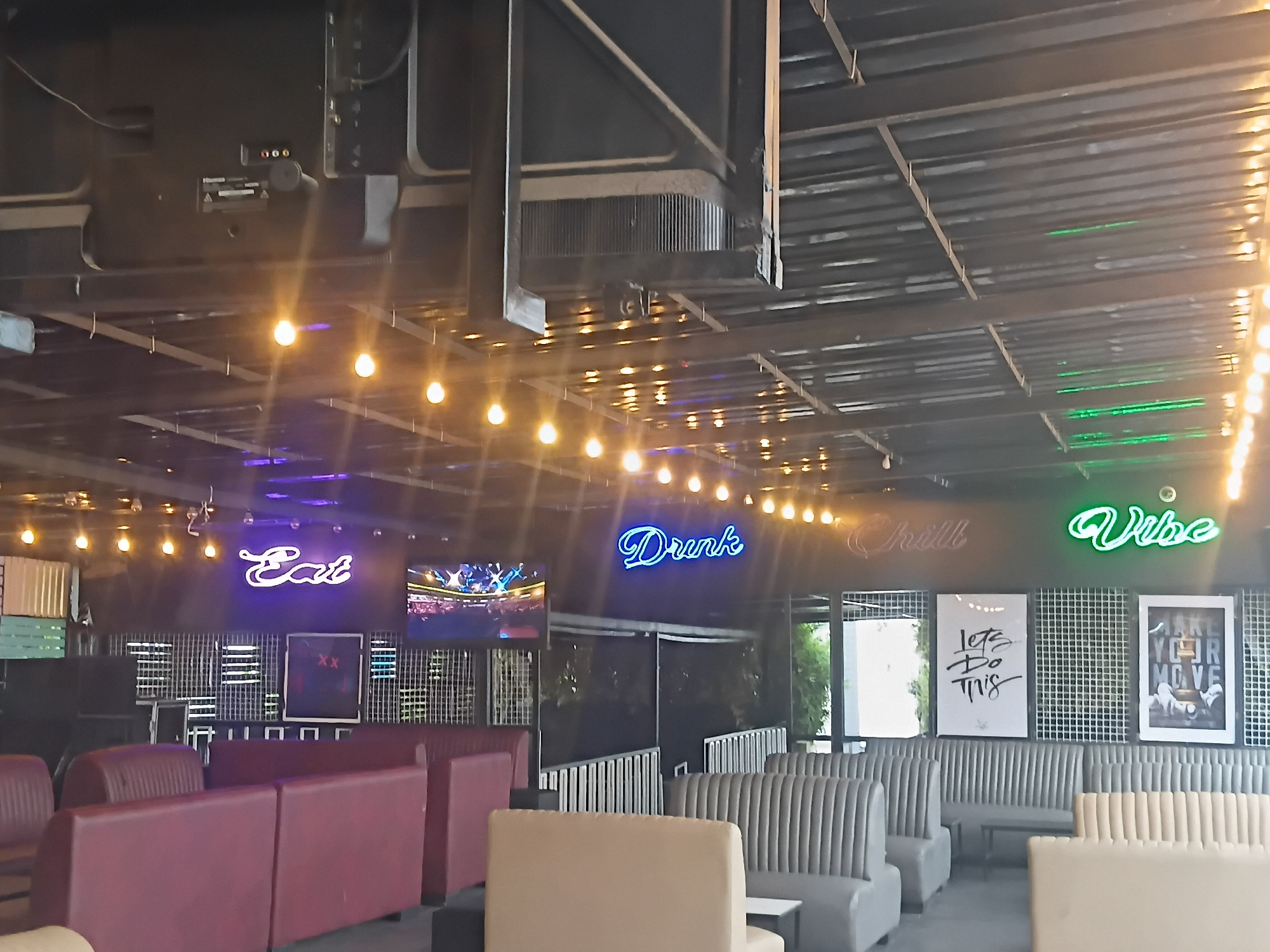 The image depicts the interior of a bar or lounge, potentially in Gura Top, Nigeria. The space is characterized by a modern industrial aesthetic with exposed ceiling beams and metal accents. Neon signs reading "Eat," "Drink," "Chill," and "Vibe" adorn the walls. There are several seating areas with booth-style arrangements upholstered in maroon, gray, and beige. Large-screen TVs are mounted on the walls, one of which displays a sporting event. The decor includes framed artwork and decorative metal mesh panels. The scene suggests a relaxed social environment intended for dining and entertainment.