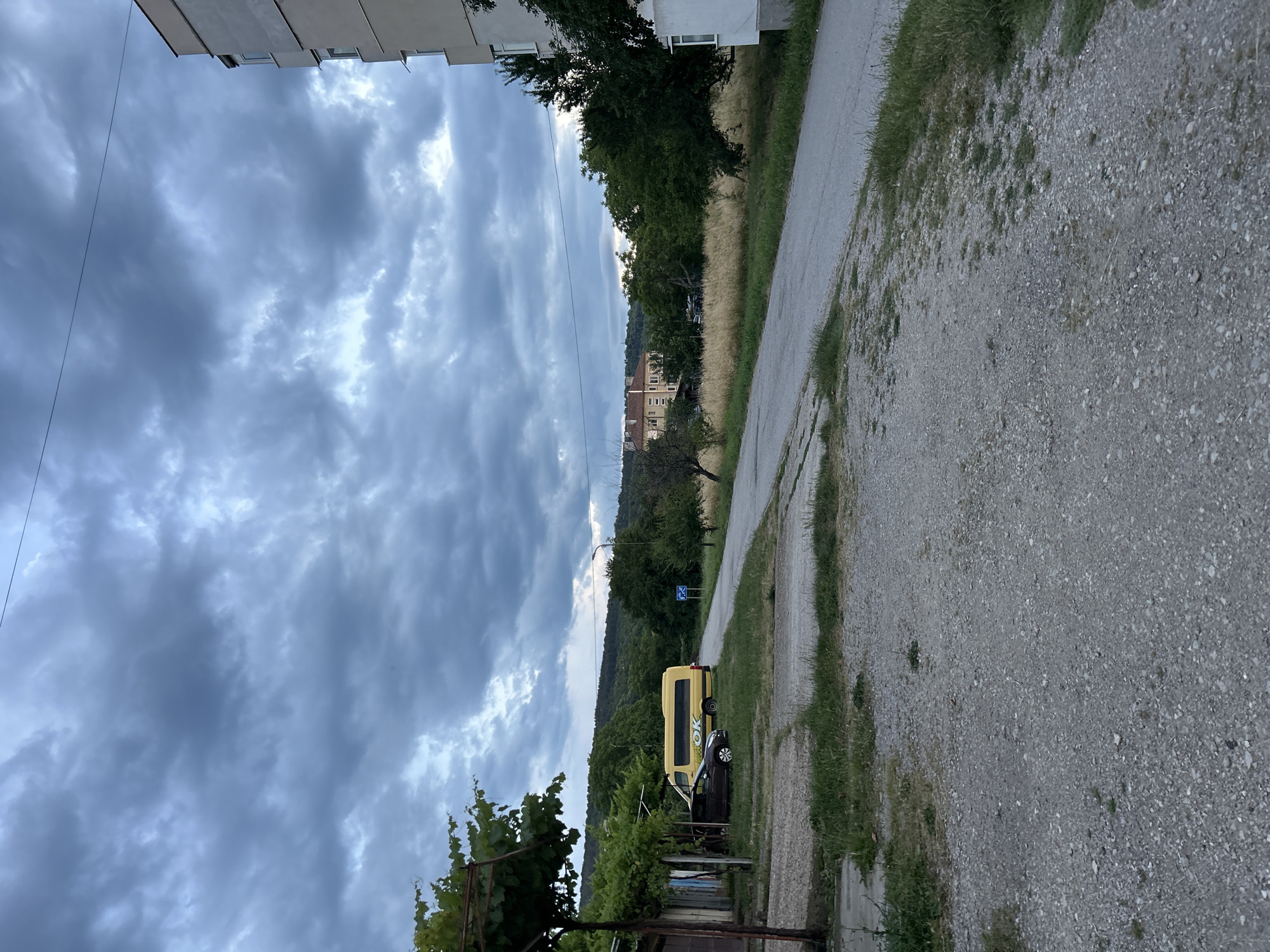 The image shows an exterior view of a street with a cloudy sky overhead. In the foreground, a gravel path leads to an asphalt road. To the left, a dark-colored car is parked next to a yellow bus with "OK" written on its side, under a vine-covered structure. A building with a light-colored facade is visible on the right side of the image. Beyond the road, there is a field of dry grass and trees, with buildings and a tree-covered hill in the background. A single streetlamp is present on the right side of the road.