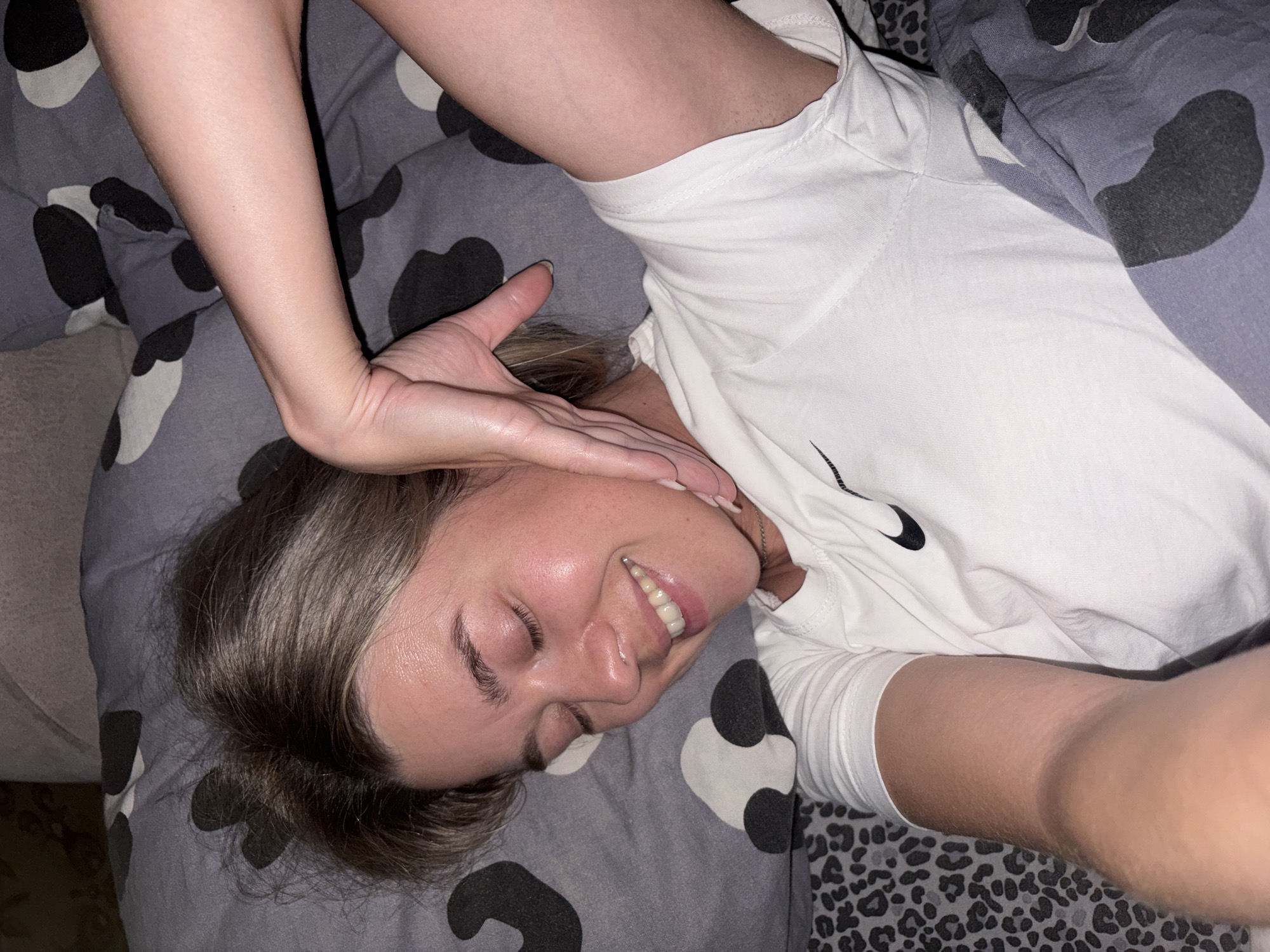 A woman with closed eyes and a smile is lying in bed. She has light brown hair and is wearing a white t-shirt with a Nike logo. Her left arm is raised, with her hand resting on her cheek. The bedding is patterned with black and grey shapes. The scene is likely indoors, in a bedroom. The location in the city of Chaikovsky, Russia, is not discernible from the image.
