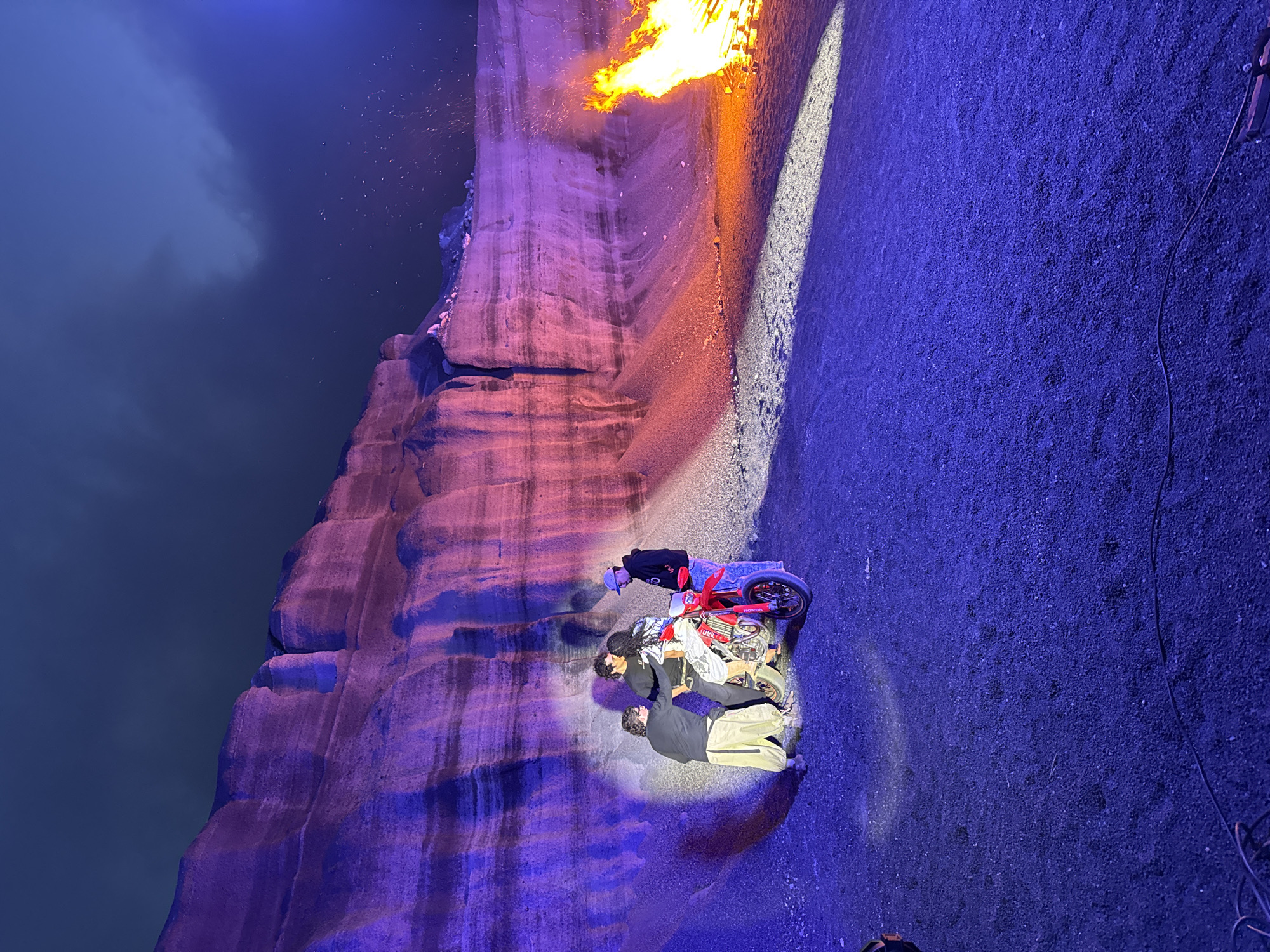 The image captures a scene in Teguise, Spain. Three individuals are positioned near a red Honda motorcycle. One person is standing next to the motorcycle. To the left, a rock wall rises, illuminated with pink and purple hues. At the top of the wall, fire is burning. The ground is illuminated with blue and white lights, with the blue light predominating. A body of water is visible in the background.