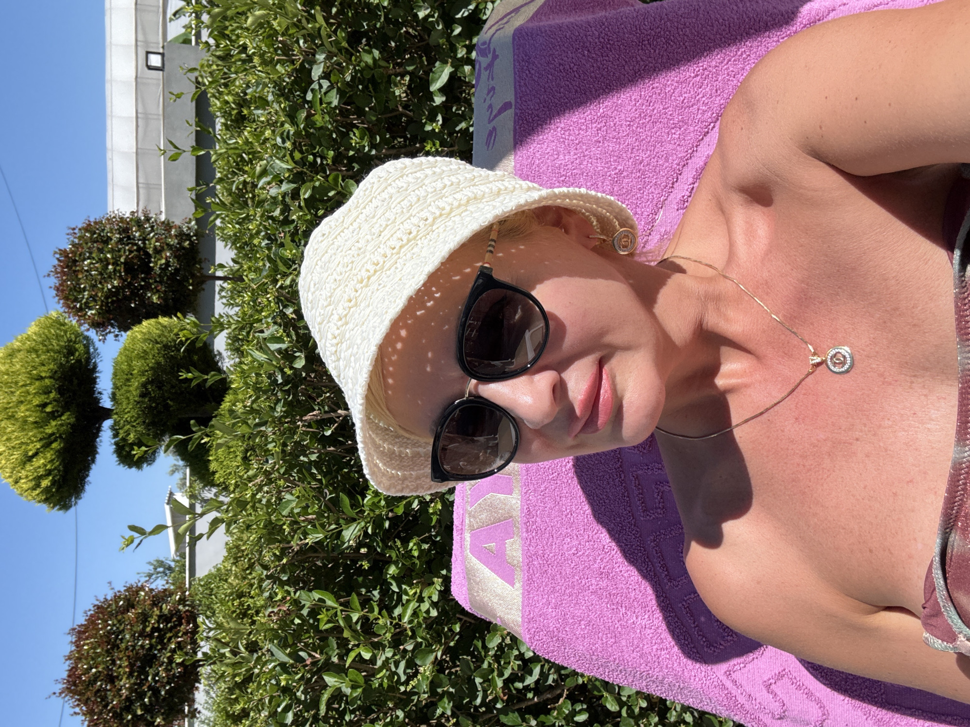 A medium shot shows a Caucasian female wearing sunglasses and a woven hat lying on a purple towel outdoors in daylight. Her head is angled slightly toward the camera, with her body primarily visible from the neck and shoulder area. She wears a delicate gold necklace. Her skin tone is reddish. The background consists of a dense green hedge and shaped trees, with a building visible in the upper left. The location is Unknown, Unknown.