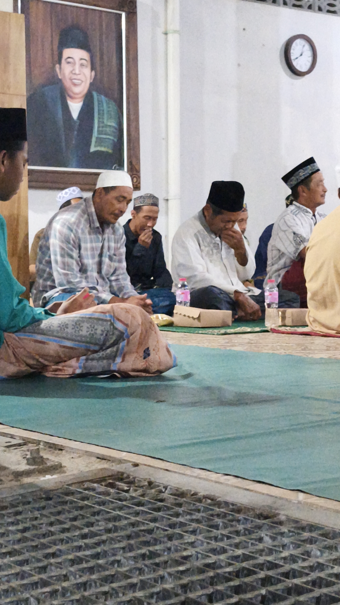 In Dalegan, Indonesia, an indoor gathering is depicted with several men seated on a green carpeted floor. A large framed portrait of a smiling man in traditional attire hangs on the wall above them. A clock is visible on the right wall. The men are dressed in modest clothing, many wearing skullcaps or turbans. Some are looking down, while others appear to be engaged in conversation or prayer. Food items wrapped in brown paper are placed in front of some individuals, along with bottles of water. The floor in the foreground features a metal grate. The overall atmosphere suggests a religious or community event.