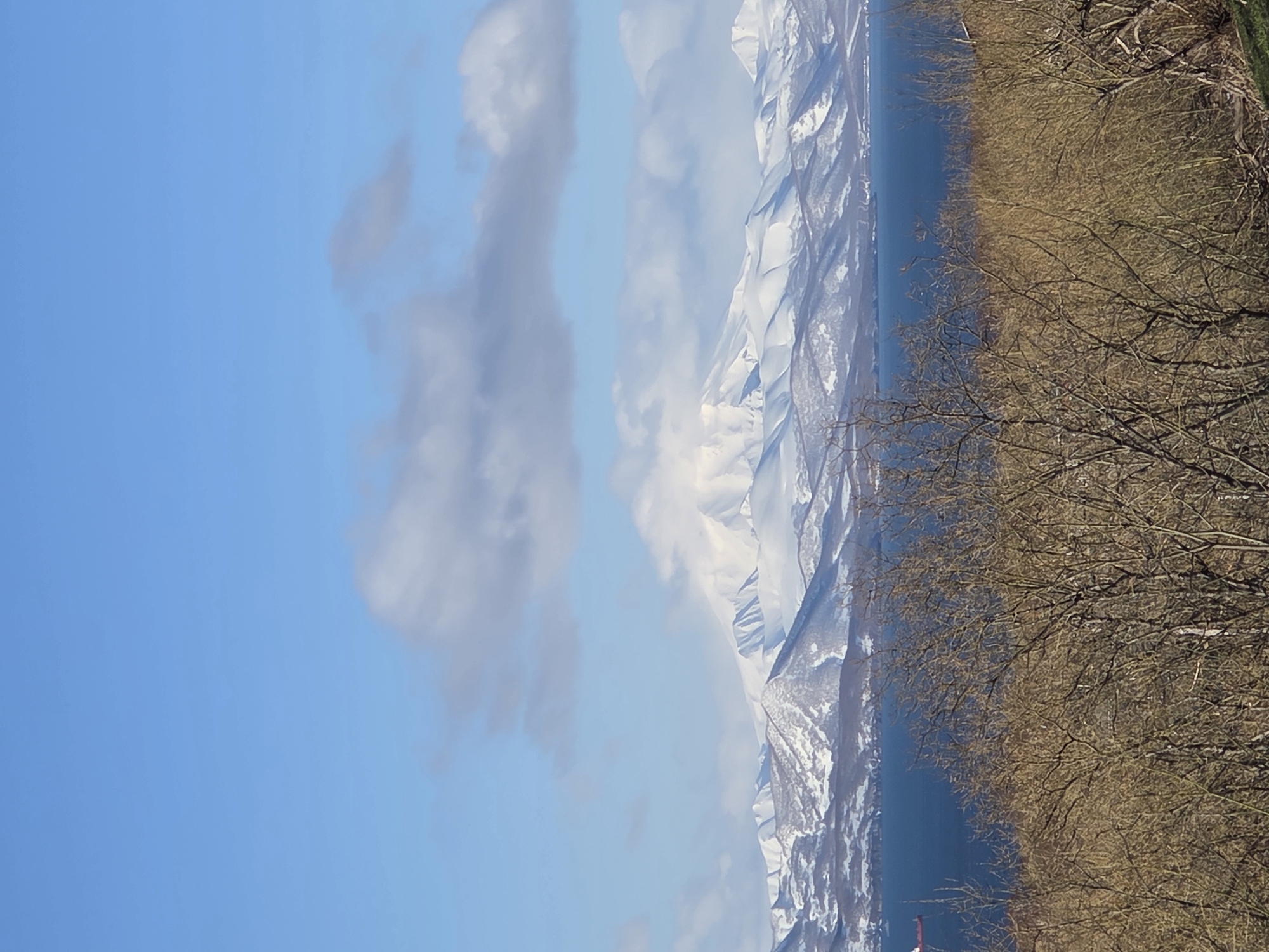 The image depicts a landscape featuring a blue sky with scattered white and gray clouds. A mountain range covered in snow and ice stretches across the frame. A body of water is visible between the mountains and a land area with bare trees. The location is unspecified.