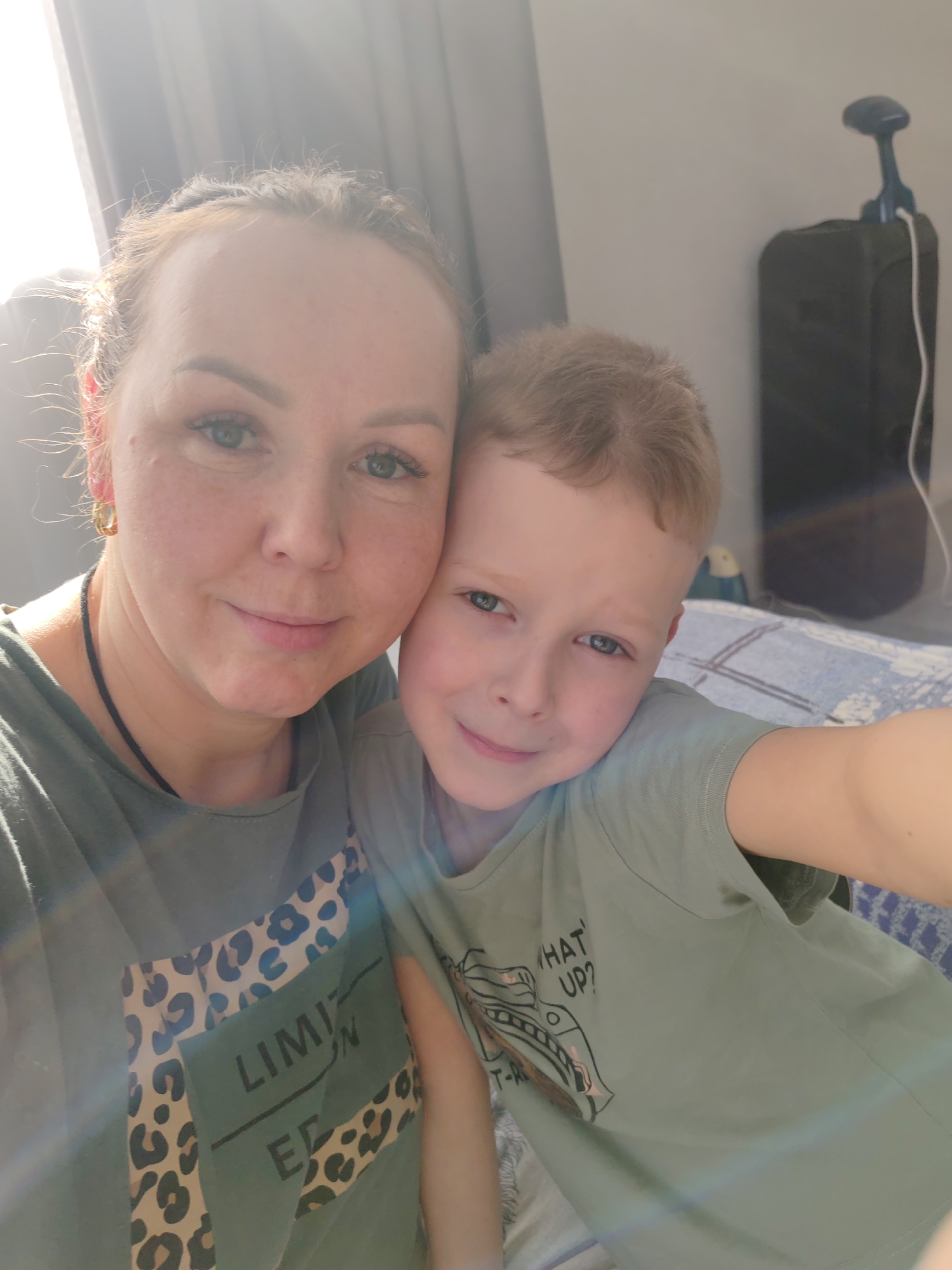 The image is an indoor selfie featuring an adult female and a young male child. The female, appearing in her 30s-40s with light skin, light brown hair pulled back, green/blue eyes, gold hoop earrings, and a black cord necklace, wears a dark olive-green t-shirt with leopard print details and "LIMIT ED" text. She holds the camera, positioned slightly to the left, and smiles faintly. The child, approximately 5-8 years old with light skin, light brown hair, and green/blue eyes, is positioned to her right, leaning against her. He wears a light olive-green t-shirt with a "WHAT UP?" graphic and smiles faintly towards the camera.
Bright natural light enters from the upper left, causing a lens flare and backlighting the subjects. The background on the left consists of a light-colored curtain or wall. The background on the right features a light grey wall. A dark, upright object, possibly a piece of luggage or a speaker, is visible on the far right, with a black, handled device and a white cable resting on top of it. Below this, a bed or sofa covered with a blue and white patterned quilt is partially visible. No specific location within the city of Chaikovsky, Russia, is discernible from the visual information provided; the scene appears to be a private indoor residence.