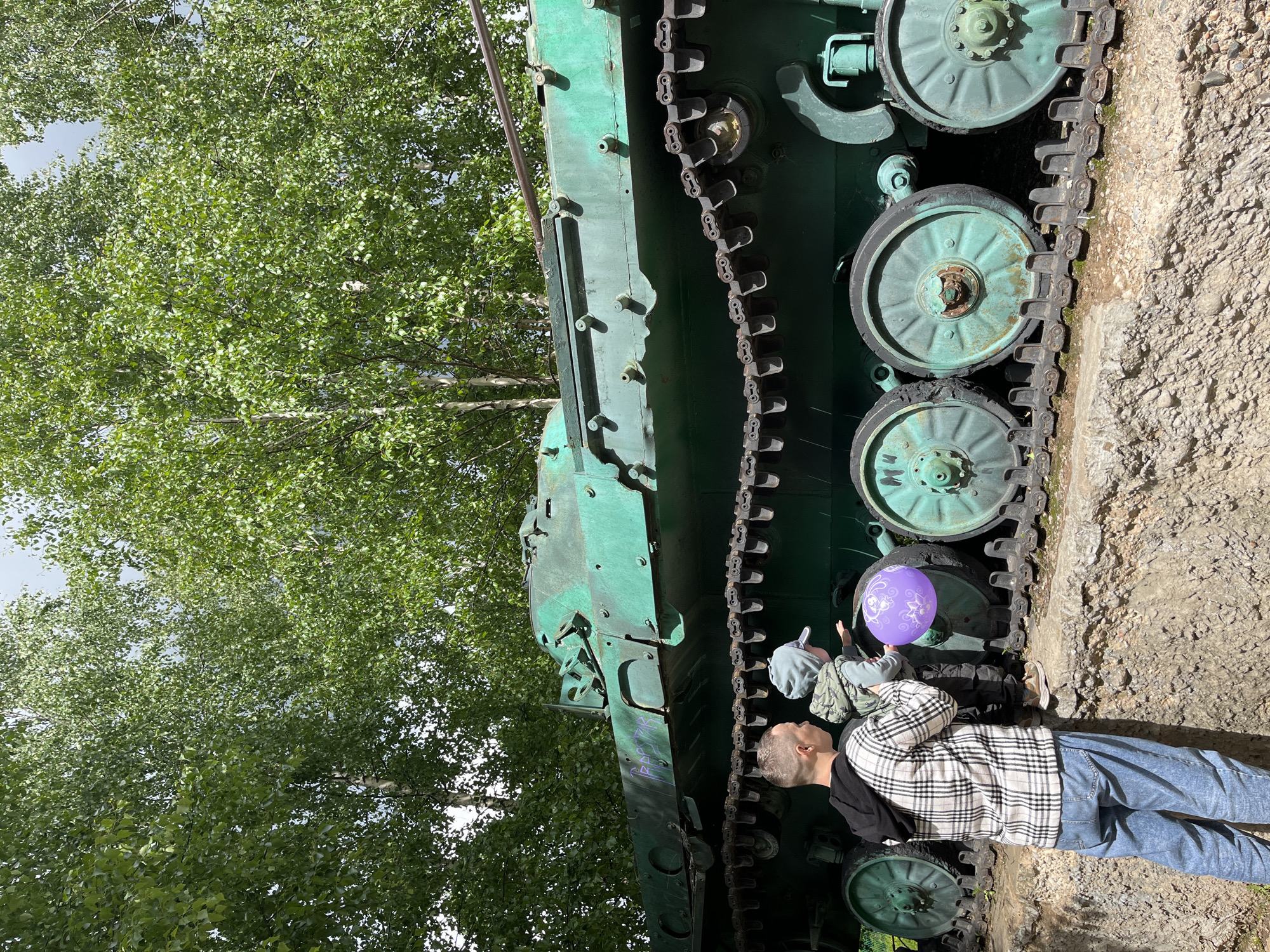 A medium-shot photograph captures two children and an adult near the tracks of a green tank in Igra, Russia. The adult, wearing a black shirt under a black and white checkered jacket and blue jeans, stands to the right, looking at the tank's tracks. To their left, a child holds a purple balloon, and another child with a gray hat looks at the tank. The tank is positioned vertically in the image, showcasing its green exterior, track wheels, and tracks. Trees with green foliage are visible in the background, contrasting with the ground made of dirt and small rocks.