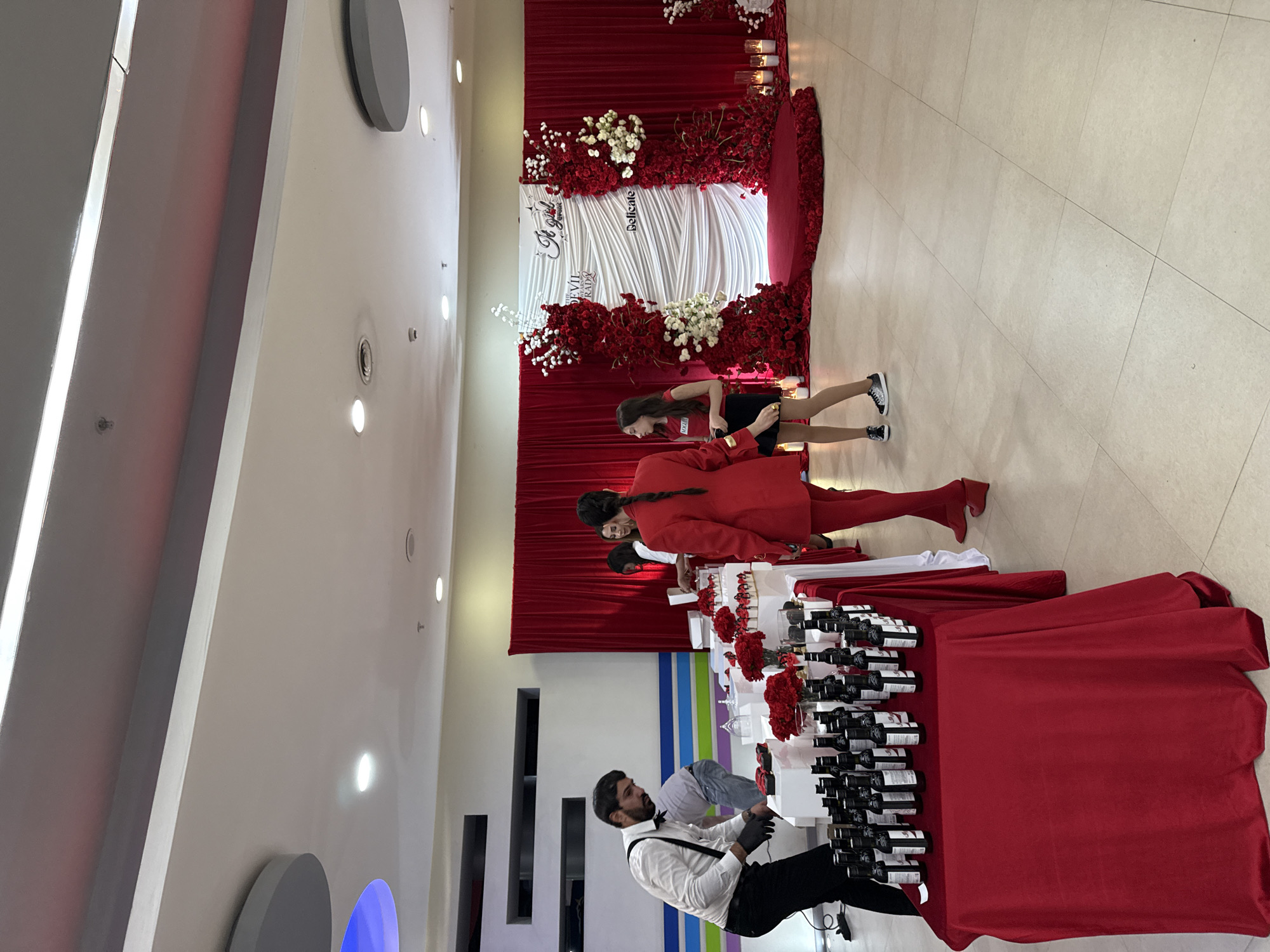 The image captures an event or gathering inside what appears to be a banquet hall or event space. In Yerevan, Armenia, a large backdrop adorned with red and white flowers is visible, along with a sign that reads "Devil Trade Delicate." In the foreground, a long table covered with a red tablecloth displays numerous wine bottles and floral decorations. A man dressed in a white shirt, black pants, and bow tie, possibly a waiter or server, is seen behind the table. Several people stand around the table, appearing to be involved in the event. A girl in a red coat, or suit, is standing by the table, and another in a red t-shirt and black skirt is walking away from the table. The floor has light-colored tiles, and the walls are a neutral shade. Above, a ceiling with recessed lighting is visible.