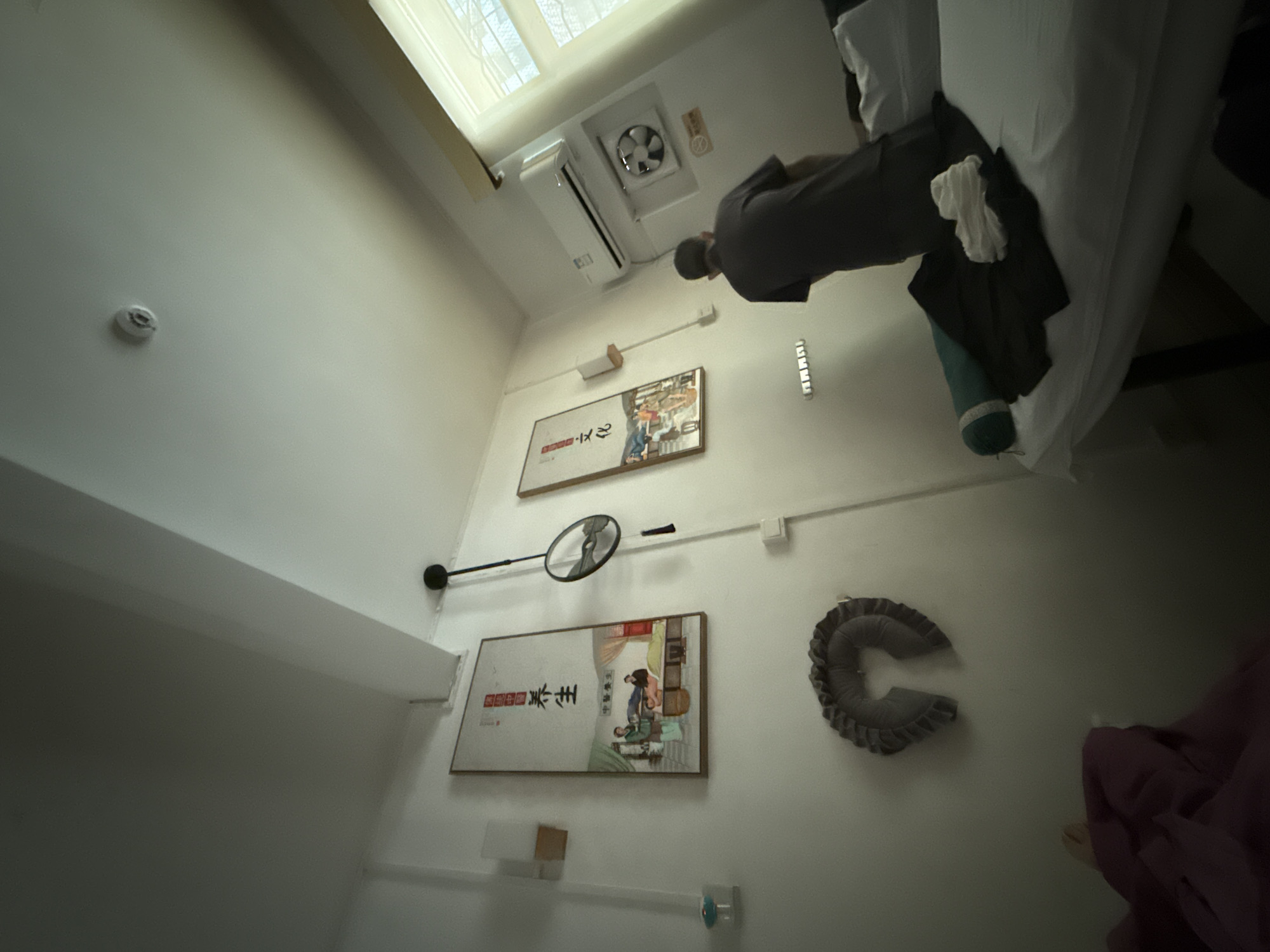 The image depicts an indoor scene, likely a massage or treatment room, in Huizhou, China. A man, seen from the back, is standing and appears to be preparing or attending to a massage table. The room is sparsely decorated with two framed artworks on the wall, featuring what appear to be traditional Chinese illustrations related to health or culture. Various objects are present, including a U-shaped neck pillow hanging on the wall, an air conditioning unit, and a fan. The lighting suggests it is daytime, with light coming in from a window on the right side of the frame. The overall impression is one of a functional and perhaps therapeutic space.
