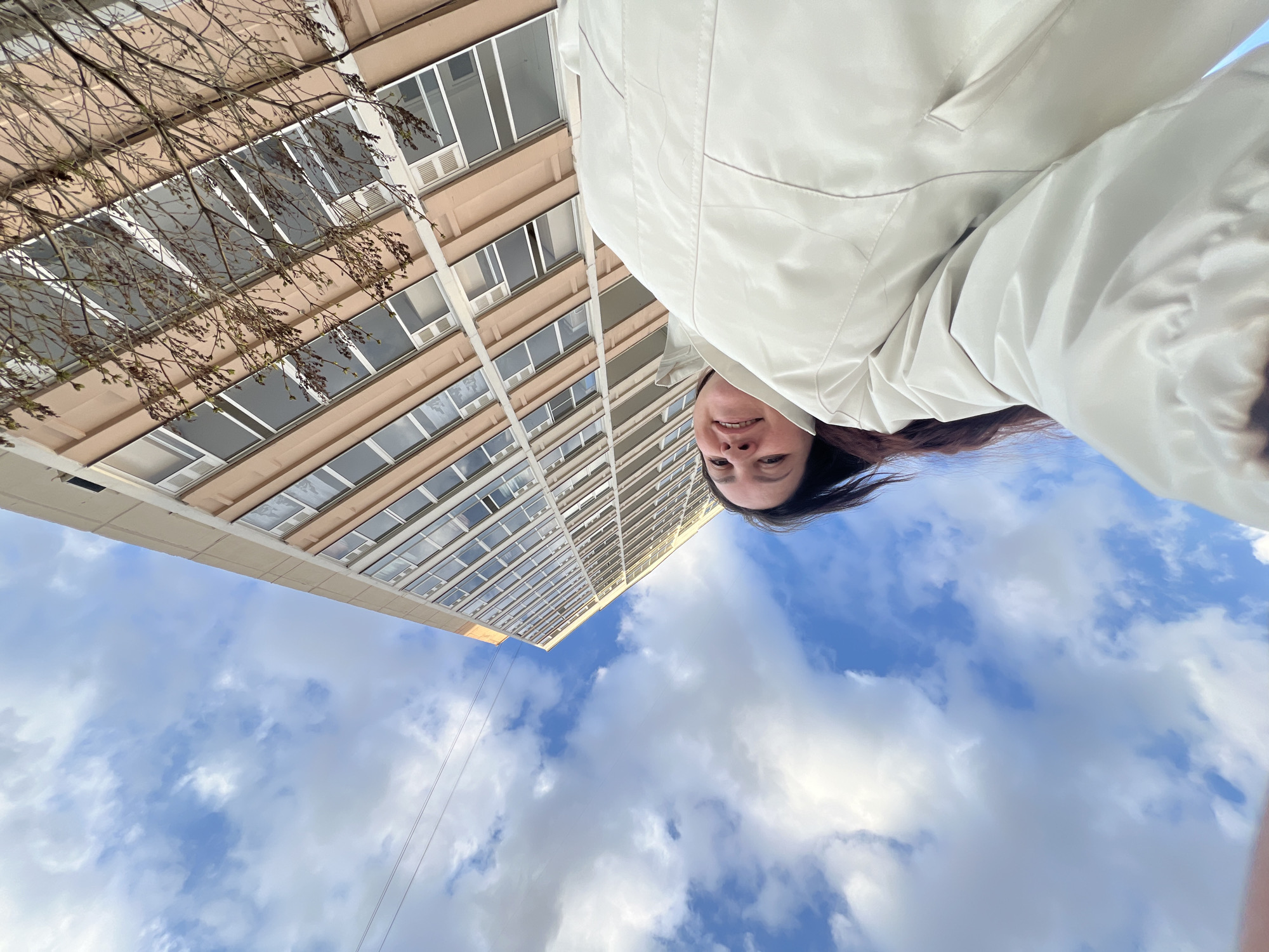 This image is an inverted, low-angle selfie, looking upwards towards a tall building and the sky. The perspective places the viewer directly beneath the subjects.
In the upper right portion of the frame, a woman is visible, appearing upside down relative to the building. Her face, from the forehead to the chin, is visible, and she has a slight smile. Her dark hair flows around her face, and she is wearing a light-colored, possibly off-white or light beige, jacket.
The background features a tall, multi-story building on the left, an architectural structure with numerous windows and light-colored (beige or light brown) facade panels. Bare tree branches, suggesting late winter or early spring, extend from the left, partially obscuring the building's lower sections. The rest of the background is dominated by a bright blue sky with scattered white, fluffy cumulus clouds. A thin wire or cable runs vertically in the lower left quadrant of the sky.
The activity depicted is a unique selfie, characterized by the unusual upward and inverted angle. The time of day is clearly daytime, given the bright illumination. The weather appears fair with partial cloud cover. Although not explicitly discernible from the image, the location is Perm, Russia, as provided by the context. There is no visible text in the image.