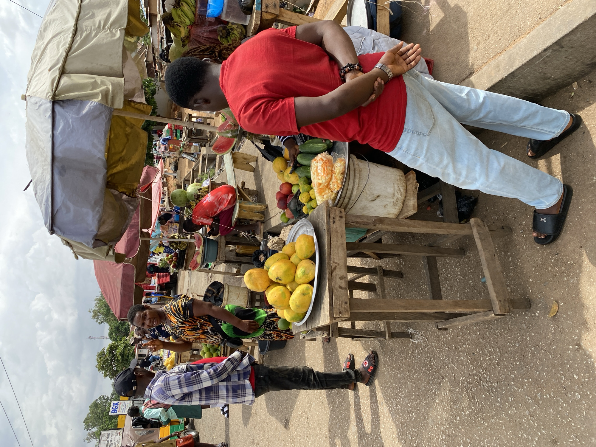 An outdoor market scene in Bukuru, Nigeria, is captured from a slightly elevated perspective and rotated counter-clockwise. The image features several people and various market stalls laden with fresh produce under an overcast sky.
In the center-right, a man wearing a red t-shirt, light blue jeans, and black slides with an 'H' logo stands with his hands clasped behind his back, looking down at a display of fruits. He has short dark hair, a beaded bracelet on one wrist, and a silver watch on the other. Slightly to his left and behind him, a woman in a black and orange patterned top smiles towards the viewer, appearing to be a vendor behind a wooden stall. In the lower-left, a man in a blue and white plaid shirt, dark pants, and a black baseball cap is partially visible, also wearing black slides with a 'BE' logo. Other figures can be seen in the bustling background, engaged in market activities.
The market stalls are constructed from rough wooden frames, some covered by large umbrellas for shade, including a prominent beige and silver one in the upper-left and a red one further back. A large silver platter holds a generous pile of yellow fruits, likely papayas or mangoes. Other visible produce includes sliced watermelon, whole watermelons, green gourds or cucumbers, and bunches of bananas. White buckets, plastic bags, and other containers are part of the market setup. The ground is a light-colored, rough concrete or paved surface, with a curb visible along the upper edge of the ground.
The activity depicts typical market interactions, with vendors presenting their goods and customers browsing. The scene is brightly lit, indicating daytime, despite the cloudy sky. In the distant background, a white and yellow sign with the text "BGK" is visible.