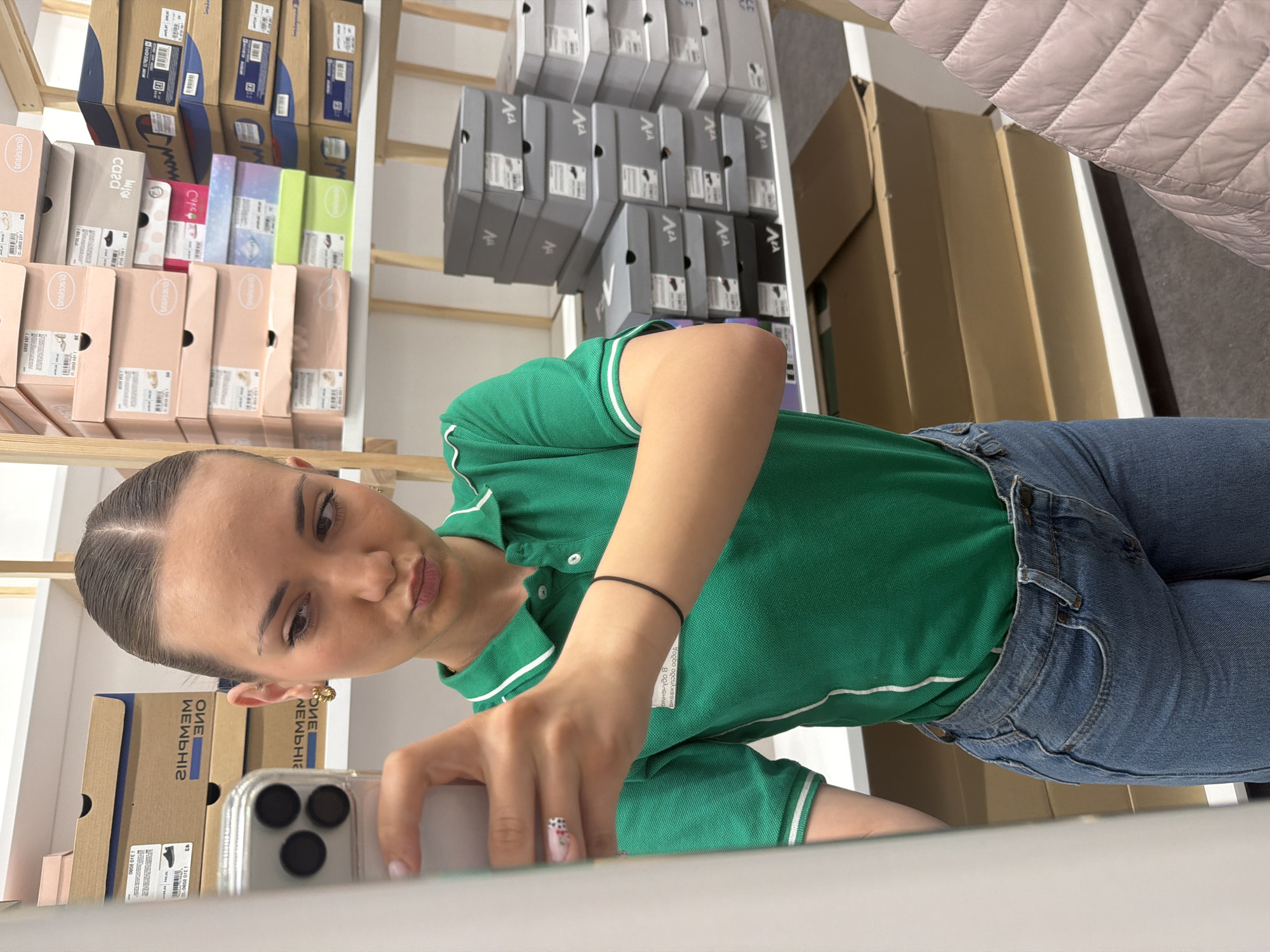 A woman is taking a selfie in what appears to be a shoe store. She is wearing a green polo shirt, jeans, and has her hair pulled back. She is holding a smartphone with multiple lenses in her right hand. Behind her, there are shelves stacked with shoe boxes, some of which are branded with the name "MEMPHIS." The shelves and the presence of shoe boxes suggest that this photo was taken in a retail environment. The image is cropped and slightly rotated, making it difficult to ascertain the exact location of the store in Sofia, Bulgaria, but the various shoe brands and box designs indicate it could be a common retail or shoe store setting. A coat is draped on top of some boxes, further indicating its retail surroundings.