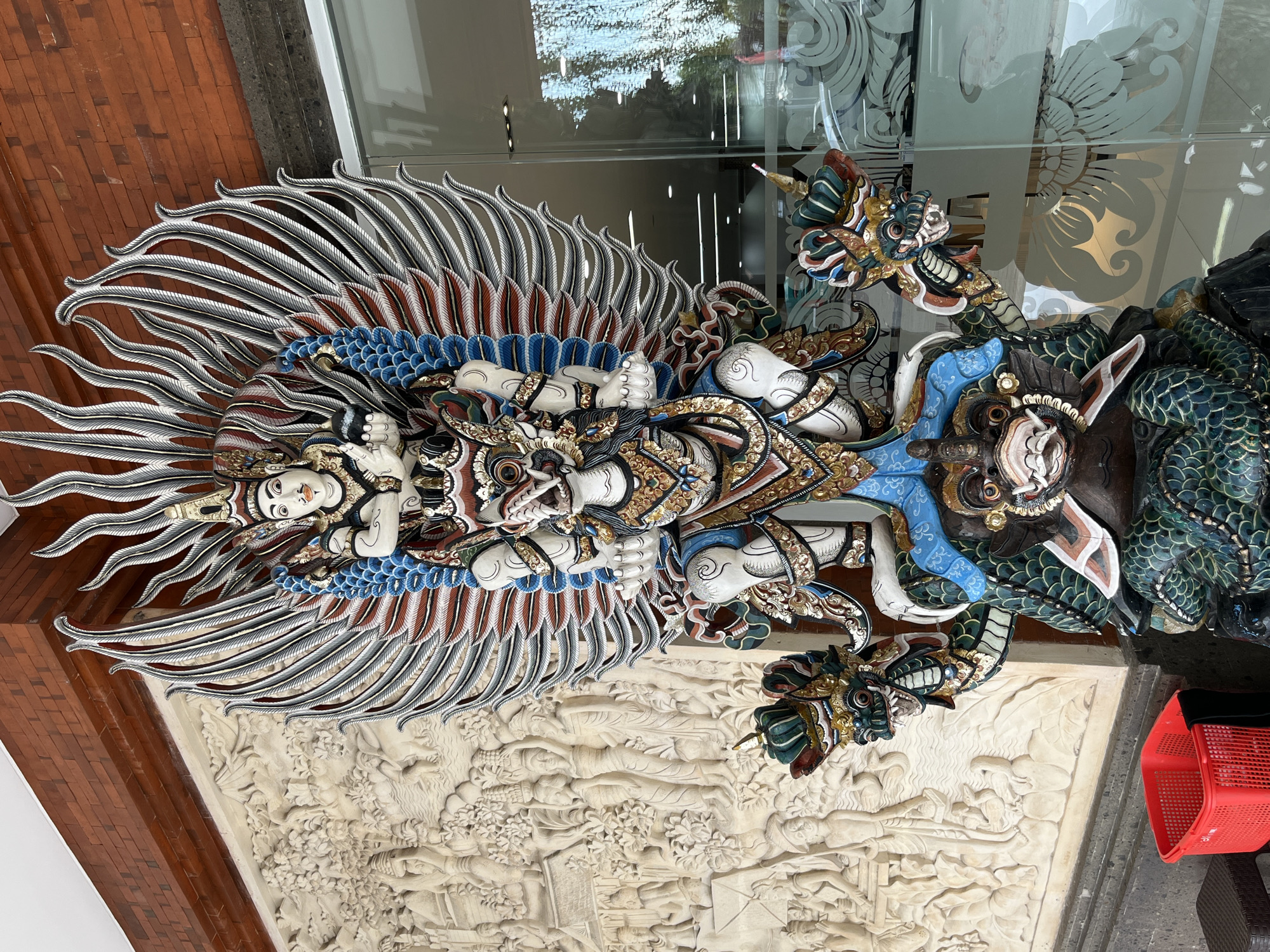 This media file showcases a vibrant, intricately carved sculpture depicting a mythical creature, likely Garuda, a celestial bird-like creature that is a mount of the Hindu deity Vishnu. The sculpture is rendered in a colorful and detailed style, with the creature's large, fanned wings prominently displayed. It appears to be made of wood and is adorned with gold accents, blue, red, and white paint. The creature has multiple arms, and its body is intertwined with what seems to be a dragon-like figure on its right.
Beneath the main sculpture is a relief carving on a lighter-colored stone, depicting a scene with several figures in a narrative style. The overall setting appears to be outdoors, possibly in a temple or an ornate entrance to a building, given the presence of brickwork and reflective glass surfaces in the background. The lighting suggests it could be daytime.
On the right side of the frame, a bright red plastic basket is visible, adding a contemporary element to the scene and contrasting with the traditional artistry of the sculpture. The style of the carving and the motifs are characteristic of Balinese art, suggesting a location in Bali, Indonesia. The presence of decorative etched patterns on the glass further enhances the artistic ambiance. There is no visible text in the image.