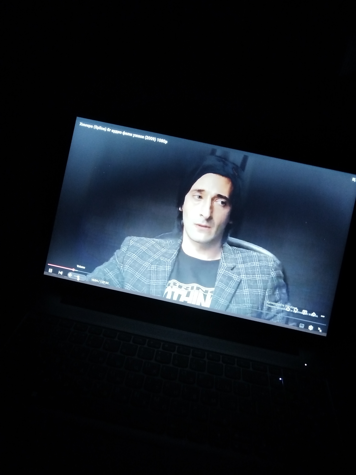 The image shows a laptop screen in a dark room, displaying the film "Chimera (Splice) бг аудио филм ужаси (2009) 1080р". Actor Adrien Brody is visible on the screen, wearing a black beanie, a t-shirt with white print, and a gray plaid blazer. The screen also displays playback controls, including "Volume" and a progress bar showing "10:04/1:47:34". The Windows operating system activation message is visible in the lower right corner. The laptop keyboard is partially visible.