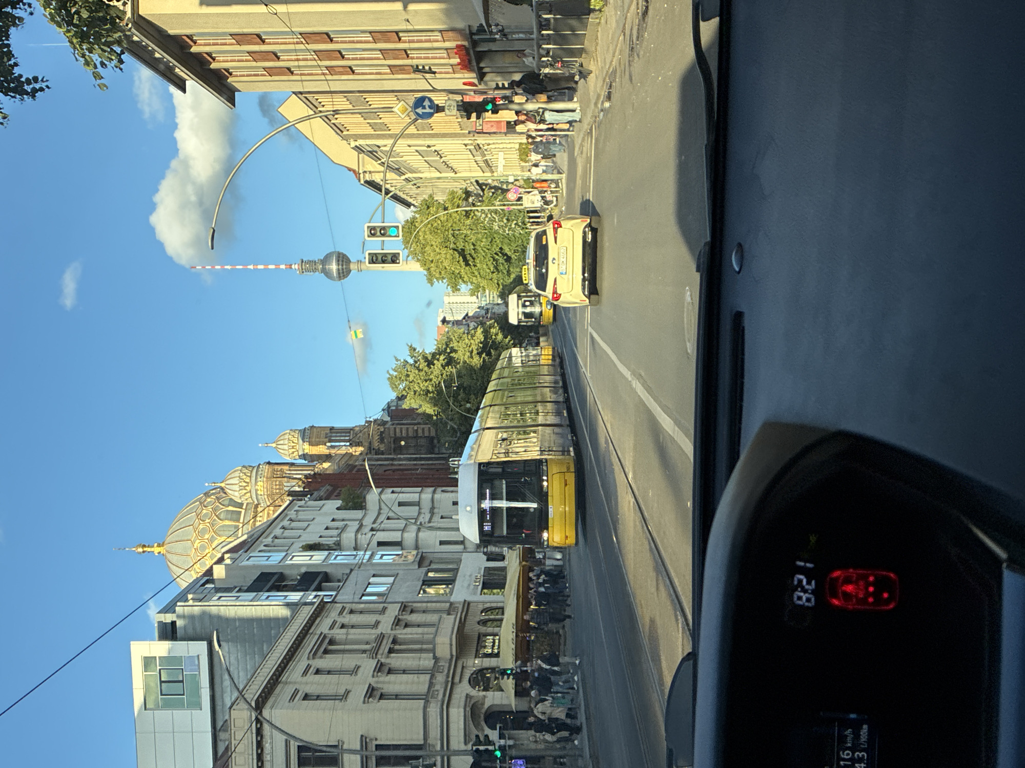 The image shows a street view in Berlin, Germany, likely taken from inside a car. In the foreground, the dashboard of the car is visible, with a digital clock displaying "8:21" and a fuel consumption indicator. The street is paved with tram tracks. A yellow tram is moving away from the camera, and a yellow taxi is also traveling in the same direction. On the left side of the street, there are buildings with shops and pedestrians. On the right side, more buildings line the street, and there are also pedestrians and a bicycle. In the background, a distinctive dome structure, possibly a religious building like the Neue Synagoge, is visible. Further in the distance, the Fernsehturm (Television Tower) is a prominent landmark against the blue sky with scattered clouds. The lighting suggests it is daytime, possibly morning or late afternoon.