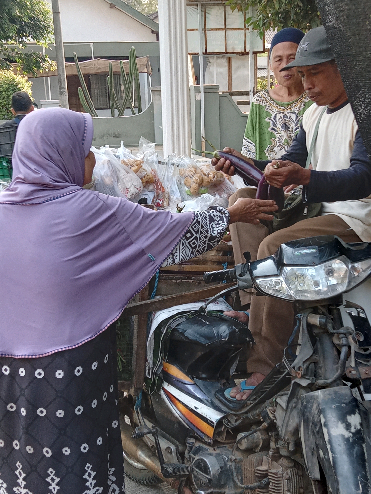 The image depicts a street scene in Sukoanyar, Indonesia, where a vendor is selling eggplants from a motorcycle. Three people are visible: a woman in a purple hijab and patterned skirt is on the left, facing away from the camera; a man in a green patterned shirt and blue head covering is in the middle, looking towards the man on the right; and a man in a white shirt and dark baseball cap is on the right, seated on the motorcycle and holding two eggplants. He appears to be interacting with the woman. The motorcycle, a common mode of transport and vendor stall in the region, is parked on a street. Bags of produce are piled behind the people, suggesting a marketplace setting. The background shows a building with a white facade and arched windows, and some vegetation. The lighting suggests it is daytime, likely morning or late afternoon.