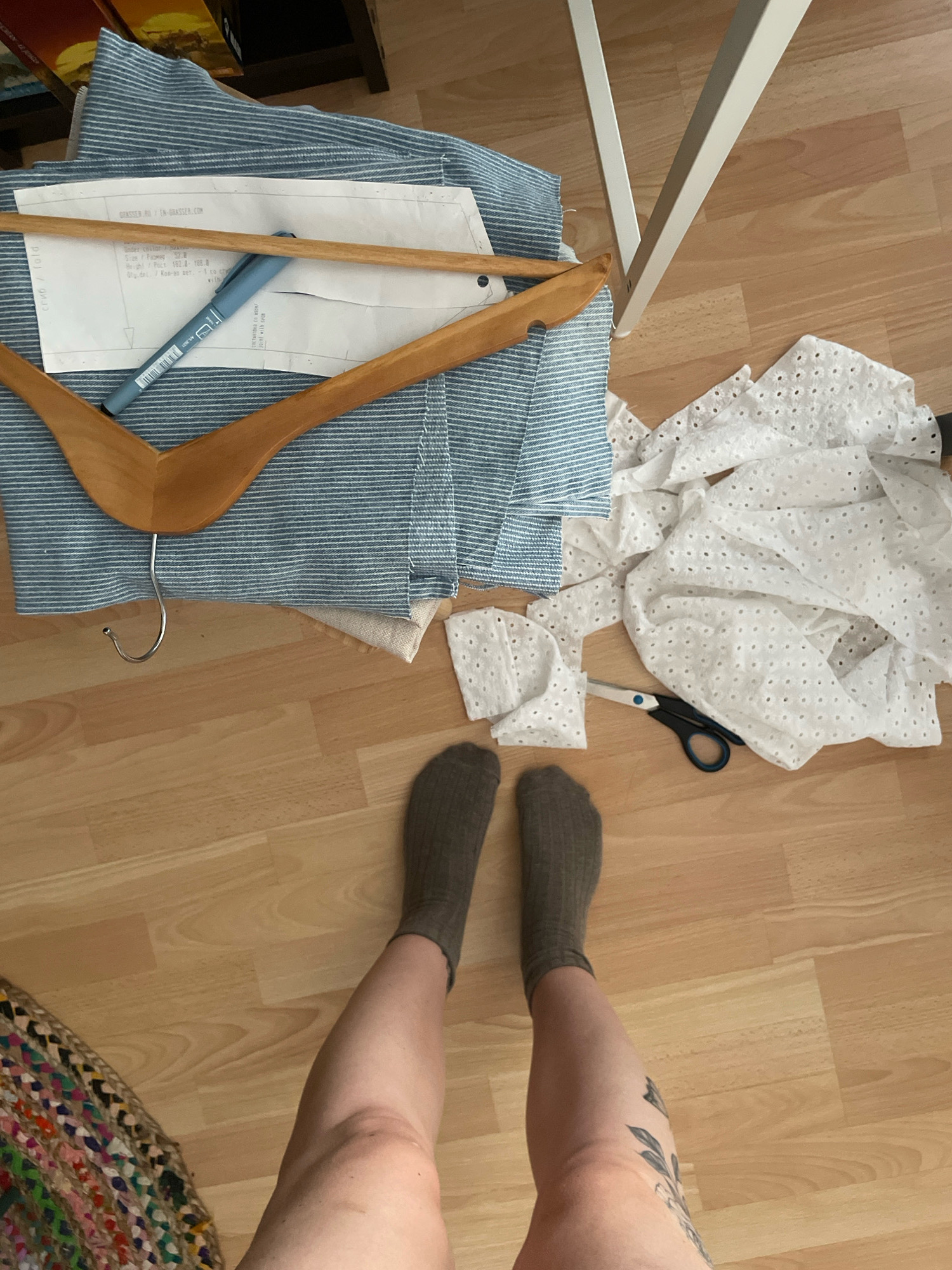 The image depicts a domestic scene on a wooden floor. In the upper left quadrant, fabric with blue and white stripes is folded, partially obscured by a wooden clothes hanger. A sewing pattern and a blue marker pen are laid on top of the fabric. To the right of this, a pile of white eyelet fabric is scattered, with a pair of black-handled scissors lying on it. In the lower half of the image, a person's legs, wearing grey socks, are visible, along with part of a colorful woven rug on the far left. The background shows the legs of a white table. There is no indication of the city of Atarfe, Spain, or any specific urban environment. The scene appears to be a DIY sewing project in progress.