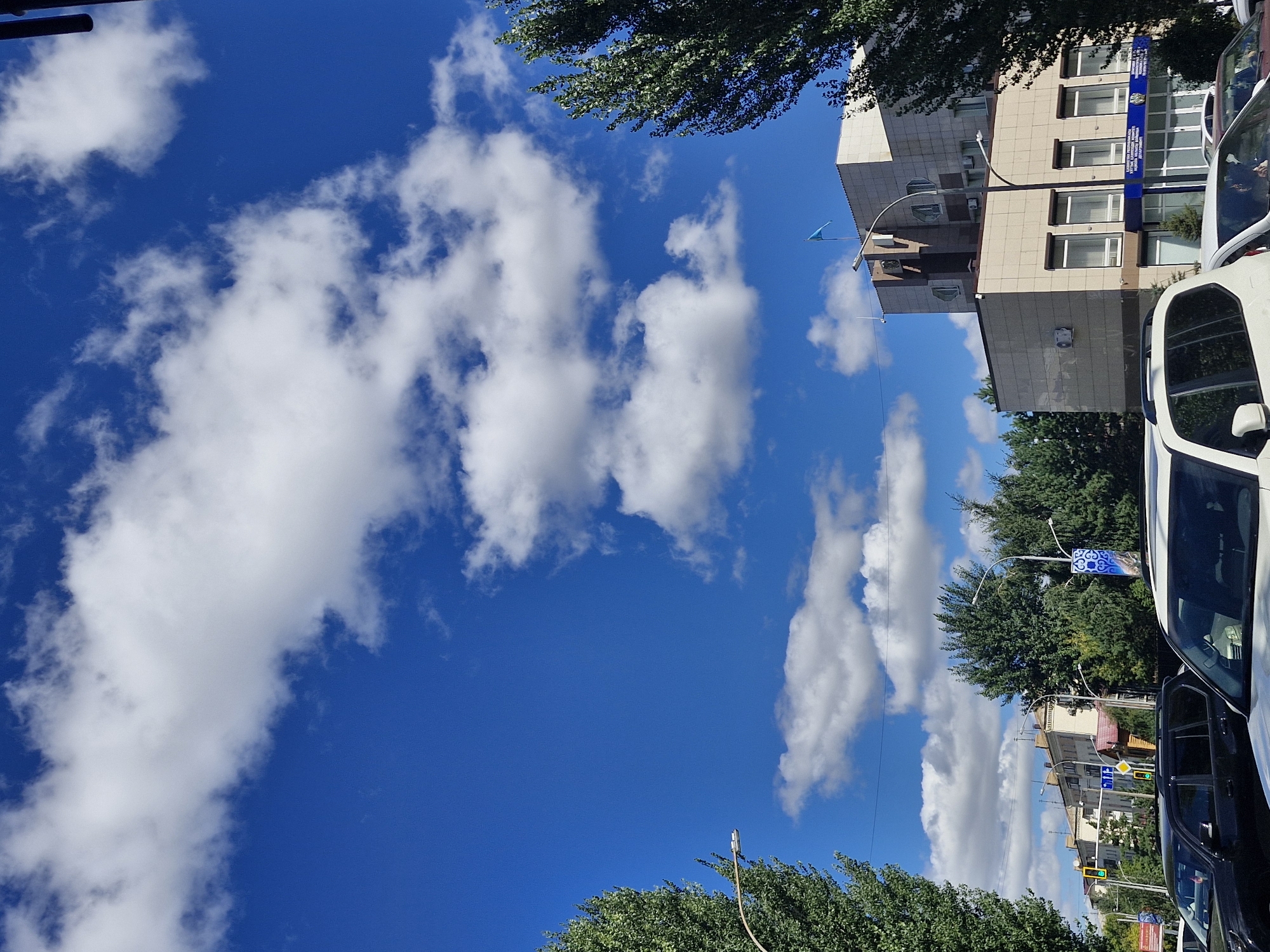 The image shows a street scene in Kokshetau, Kazakhstan, on a sunny day with a bright blue sky dotted with white clouds. The foreground is occupied by several vehicles, including a black SUV and a white SUV. In the background, there are buildings and trees lining the street. One building, likely an administrative building, is prominent, featuring a light-colored facade with windows and a flag at the top. Traffic lights and streetlights are also visible. The overall impression is a typical urban environment under a clear sky.