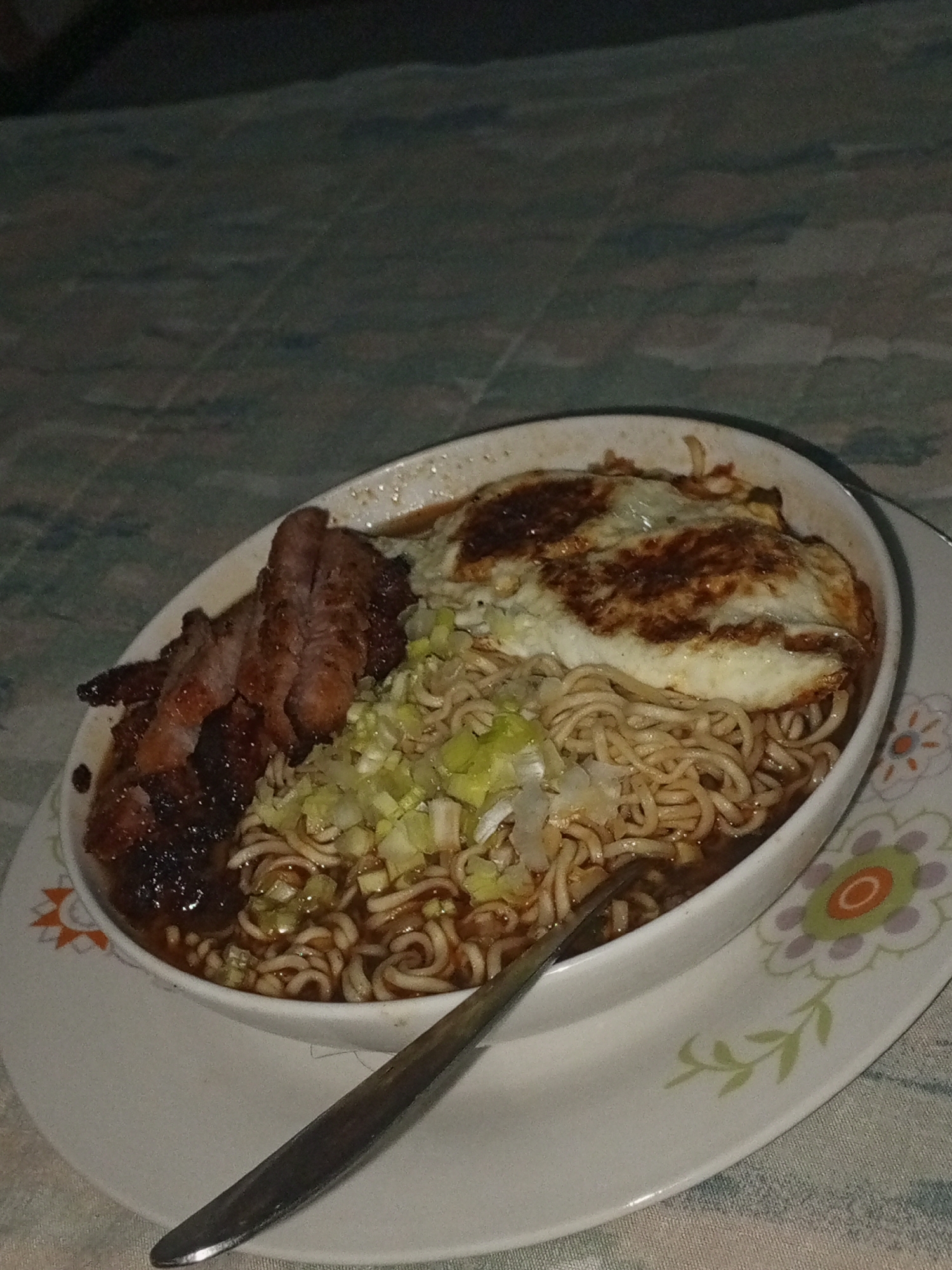 A close-up, eye-level shot captures a bowl of noodle soup with various toppings, presented on a round plate and resting on a patterned tablecloth. The scene is dimly lit, suggesting an evening or nighttime setting in an indoor environment, likely in Porlamar, Venezuela.
The central subject is a white, oval-shaped bowl filled with a dark-broth noodle soup. Visible are light-brown, wavy noodles submerged in the broth. Generous toppings enhance the dish: a pan-fried egg, with its white cooked and the edges slightly crisped and browned, sits atop the noodles. Adjacent to the egg are several thick slices of dark, roasted or grilled meat, appearing rich and somewhat caramelized. A large quantity of freshly chopped green onions is sprinkled over the noodles, adding a bright green contrast to the darker elements.
A metallic spoon is positioned inside the bowl, resting against the noodles, indicating the meal is ready to be eaten. The bowl sits on a white ceramic plate, which features a decorative floral pattern along its rim. This design includes stylized flowers with green leaves and centers in shades of orange and light purple. The tablecloth beneath has a subtle, light-colored patterned design. There is no visible text in the image.