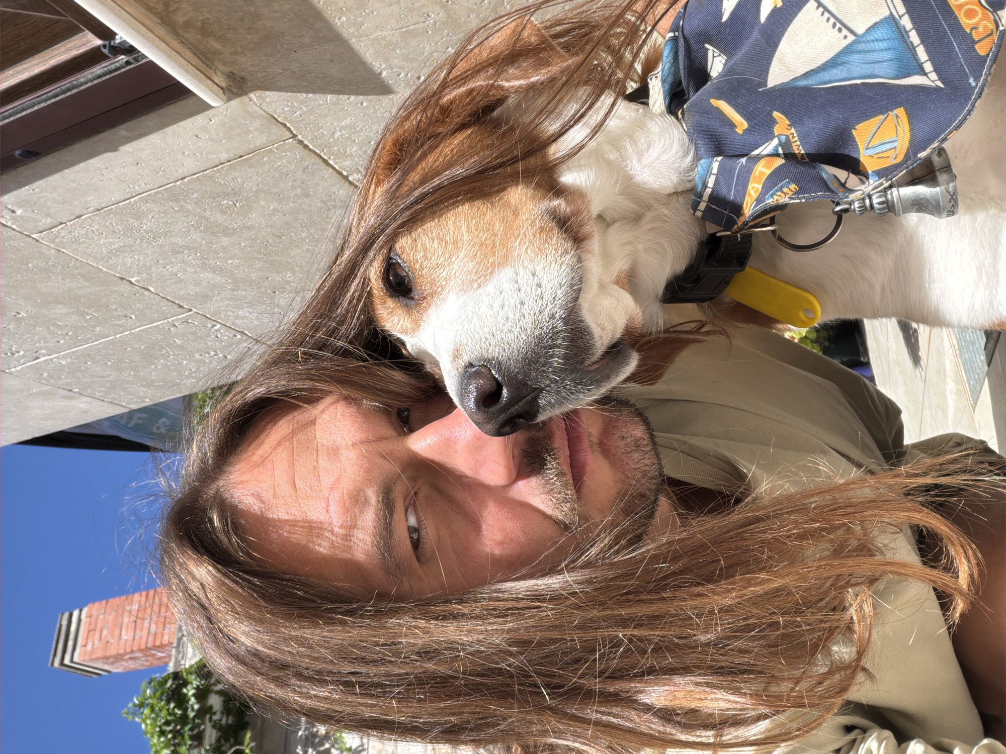 The image shows a man and a dog in Petrovac centar, Montenegro. The man has long brown hair that partially covers his face, and he is looking at the camera. The dog is a beagle, with a brown and white coat, and is lying on top of the man's head. The dog is wearing a blue and white patterned collar with a yellow tag attached. The background shows a light-colored stone surface, possibly a street or sidewalk, and a partial view of a blue sky and a brick building with green vegetation.