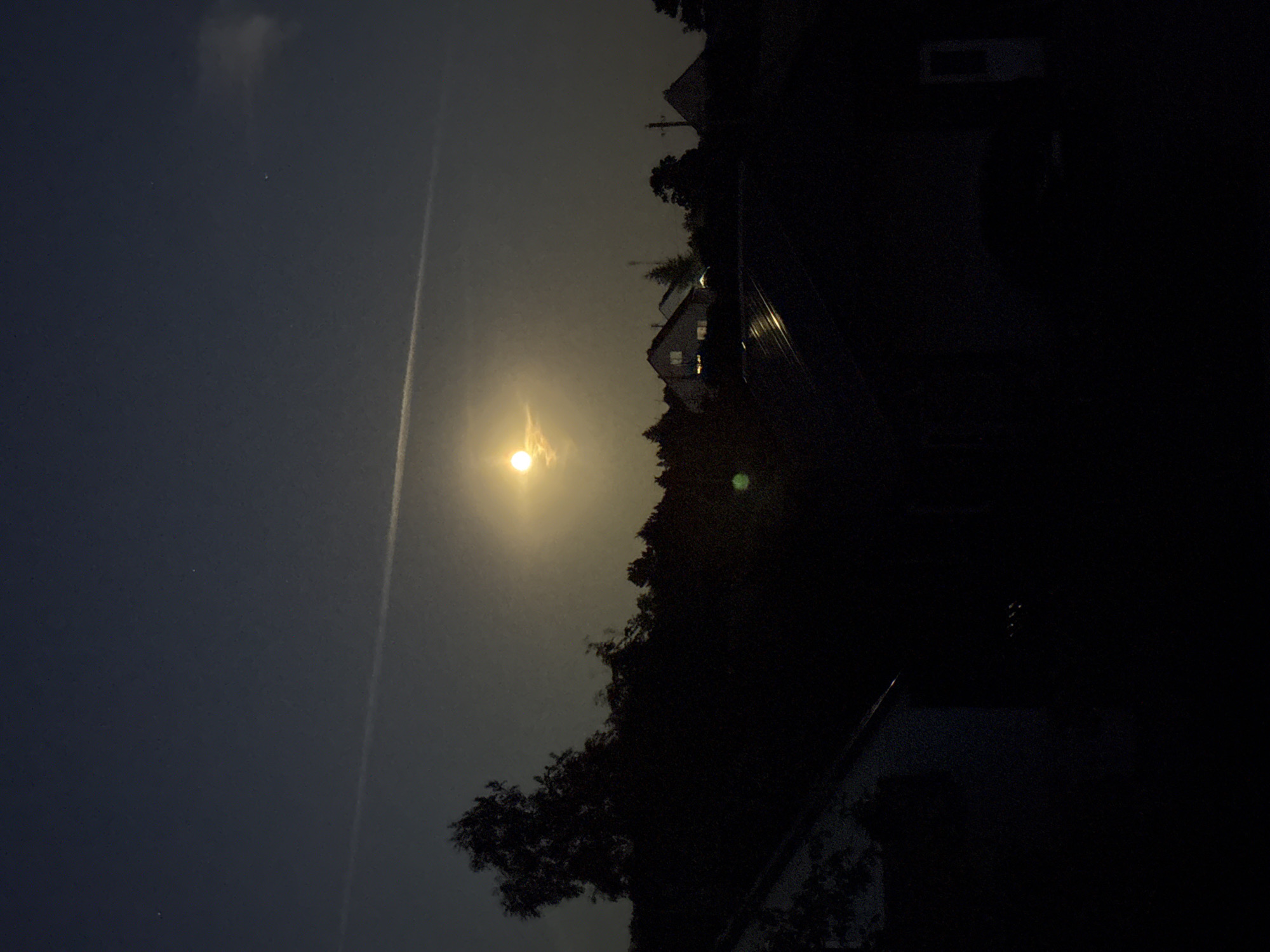 A full moon shines brightly in a dark night sky over Vikraičiai, Lithuania. A faint contrail stretches across the sky above the moon, and two distant stars are visible to the left and right of the moon. In the foreground, silhouetted trees and houses create a dark landscape. One house, in the mid-ground, has lit windows, suggesting occupancy. The moon's light illuminates the upper portion of the sky and casts a glow on the clouds around it. A green light or reflection is visible in the lower left quadrant of the image. The overall scene depicts a tranquil night in a rural or suburban setting.