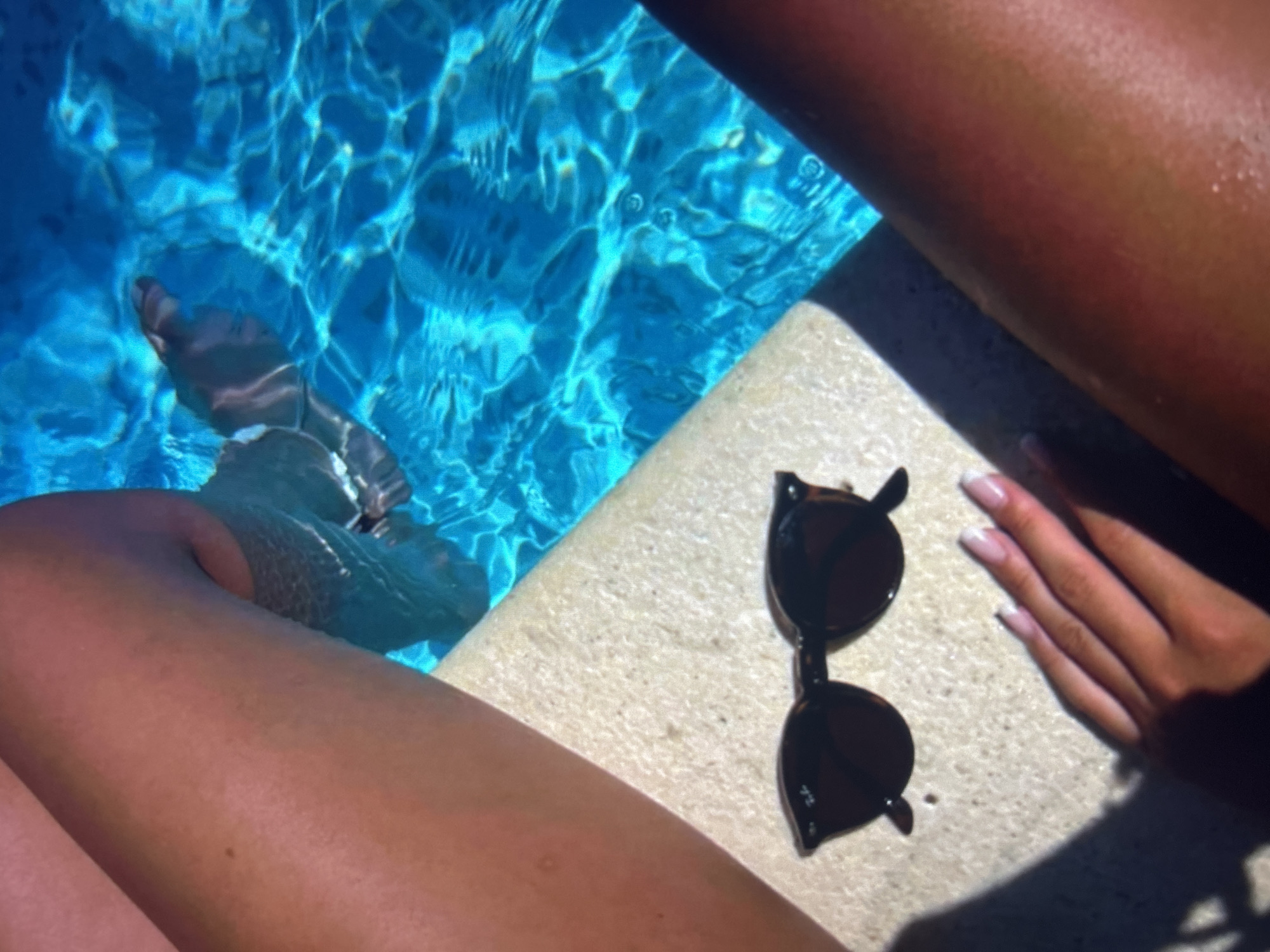 The image depicts a close-up, top-down view of a person's legs resting on the edge of a swimming pool. The pool water is a vibrant blue and appears to be illuminated by sunlight, creating ripples and reflections. On the pool's edge, in the foreground, a pair of dark-rimmed sunglasses rests on a textured, beige surface. To the right of the sunglasses, a hand with manicured fingernails is visible, with its fingers extended and slightly submerged in the water. The person's tanned legs are prominent in the frame, with one leg extending into the pool and the other positioned on the edge. The scene suggests a relaxed, summer atmosphere, likely a personal or public swimming pool. There is no visible indication of the city of Knezha, Bulgaria, within the image.