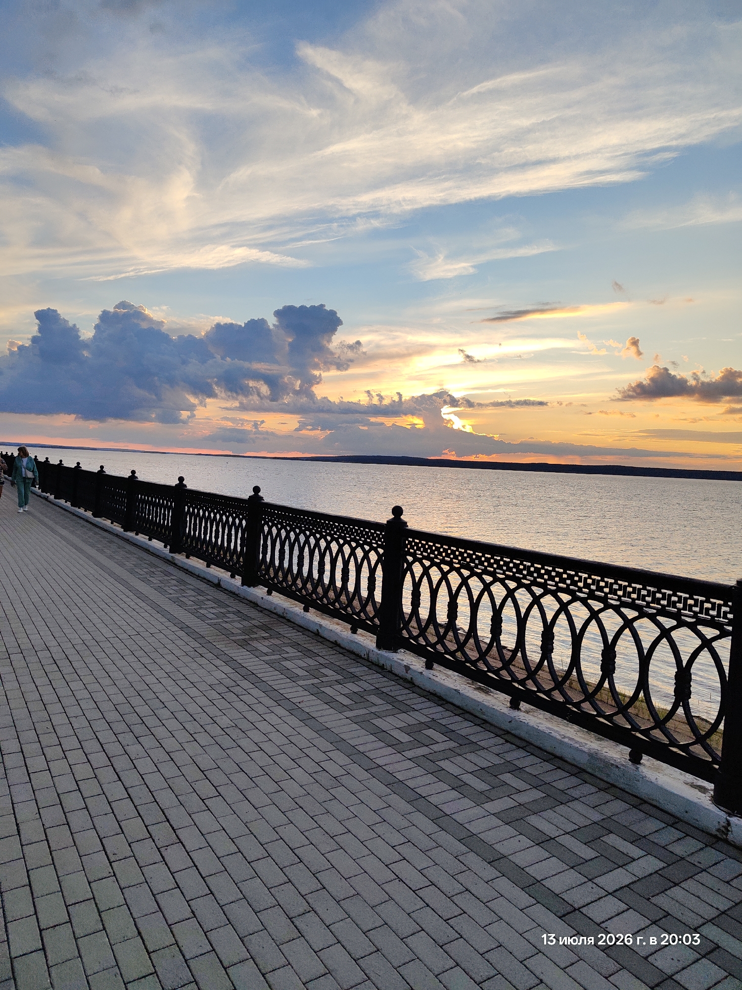 The image shows a paved promenade along a body of water at sunset. A decorative black metal railing lines the edge of the promenade, separating it from the water. The sky is filled with dramatic clouds, painted in hues of orange, yellow, and blue from the setting sun. In the distance, the horizon is marked by a faint landmass. A few people are visible walking on the promenade. The paving stones are laid in a patterned arrangement. A timestamp in the lower right corner reads "13 июля 2026 г. в 20:03".