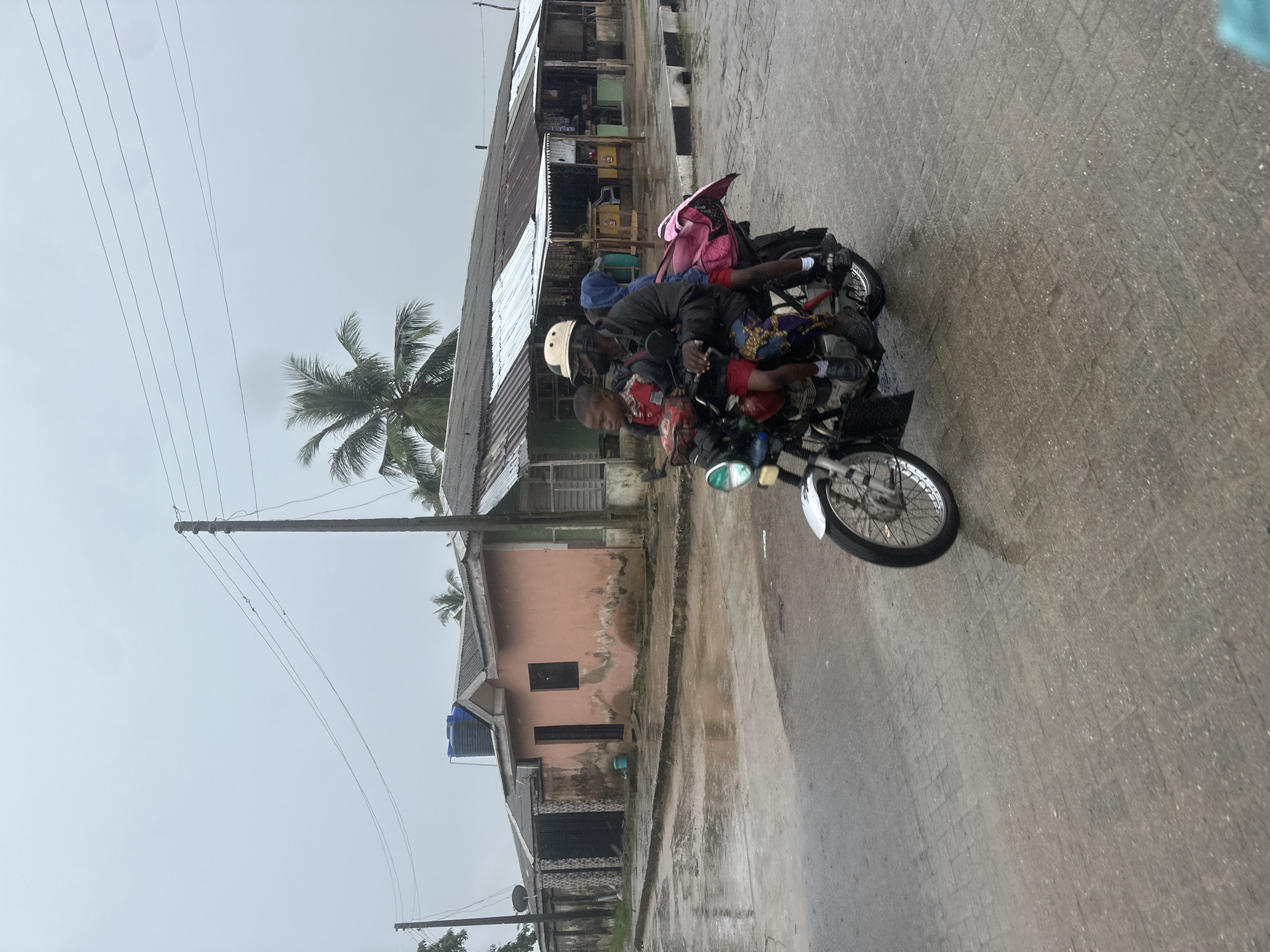 The image shows a motorcycle carrying several people on a wet, paved street. The motorcycle has a man driving, a woman behind him, and at least two children in front of the man. The woman is wearing a pink backpack. The people are dressed casually, and the man driving is wearing a white helmet. The motorcycle is carrying various items, including what appears to be a patterned cloth bag.
The scene is outdoors, with buildings visible on the left side of the street. These buildings appear to be made of corrugated metal and concrete, with some showing signs of wear and tear. There is a palm tree visible behind the buildings, suggesting a tropical or subtropical location. Overhead, power lines are strung between poles.
The street is wet, indicating that it has recently rained or is currently raining. The sky is overcast and a dull gray, suggesting a cloudy or rainy day. The overall impression is one of everyday life in a bustling, possibly developing, urban or semi-urban environment. The focus of the image is the motorcycle and its occupants, highlighting the common practice of transporting multiple people on a single motorcycle. No visible text is present in the image.