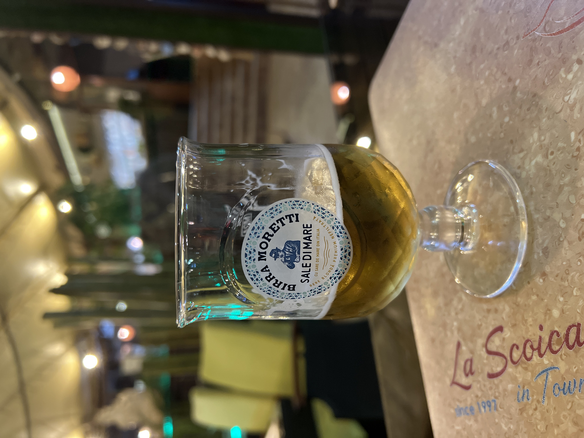 A close-up, eye-level shot captures a glass of Birra Moretti beer on a textured, light-colored surface that appears to be a table or placemat. The beer is a golden amber color and fills the lower half of a clear, stemmed glass. A white foamy head tops the beer, with some residue clinging to the inside of the glass. A circular label on the glass reads "BIRRA MORETTI SALE DI MARE" with a depiction of a man drinking. In the blurred background, there are out-of-focus elements of a restaurant or bar setting, including yellow chairs, green vertical elements that could be plants or decor, and warm lighting from overhead fixtures. The surface beneath the glass has text that is partially visible, including "La Sc..." in a cursive font, and "since 1997 in Tour...". This text suggests the location is La Scauna restaurant in Constanța, Romania, which was established in 1997. The overall scene is dimly lit, suggesting an evening or indoor setting. There are no visible people in sharp focus.