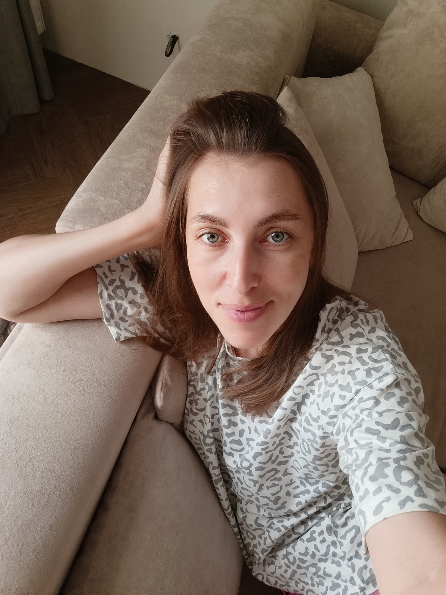 A woman with shoulder-length brown hair and blue eyes is lying on a beige sofa, taking a selfie. She is wearing a light-colored shirt with a grey leopard print. Her right arm is bent and resting on her head, with her hand touching her hair. The sofa appears to be in a living room. The flooring visible in the background is dark wood. The image was taken in the city of Chaikovsky, Russia.