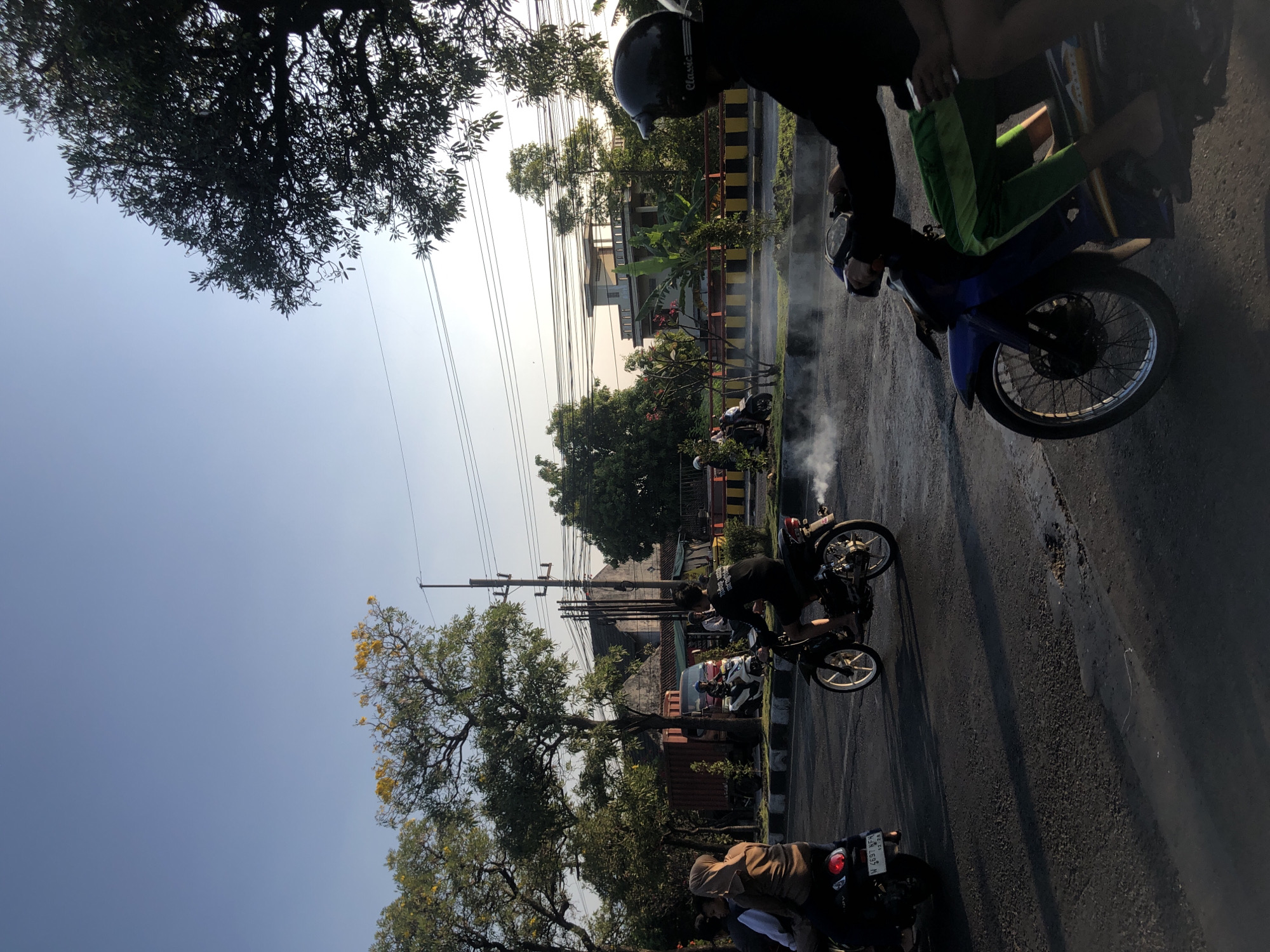 This is a medium shot, taken outdoors, of a street scene in Kludan, Indonesia. The image captures several people riding motorcycles on a paved road. The sky is clear and blue, with the sun casting long shadows across the asphalt. Trees with green foliage and some with yellow flowers line the left side of the road, with a sidewalk in between the trees and the street. In the background, there are some buildings, power lines, and more trees.
On the road, several motorcycles are visible. In the foreground, a person in a black shirt and helmet is riding a blue motorcycle, with visible smoke emanating from the exhaust pipe. Further down the road, a person in a black shirt is riding a black motorcycle with white rims, also producing smoke. To the left, two people are on a black scooter, with the passenger wearing a tan hijab and brown long-sleeved shirt. Another motorcycle, ridden by a person in a white shirt, is further back. A red truck is parked behind the sidewalk on the left.
The overall impression is a snapshot of daily life in an Indonesian city, likely during the late afternoon given the lighting and shadows.