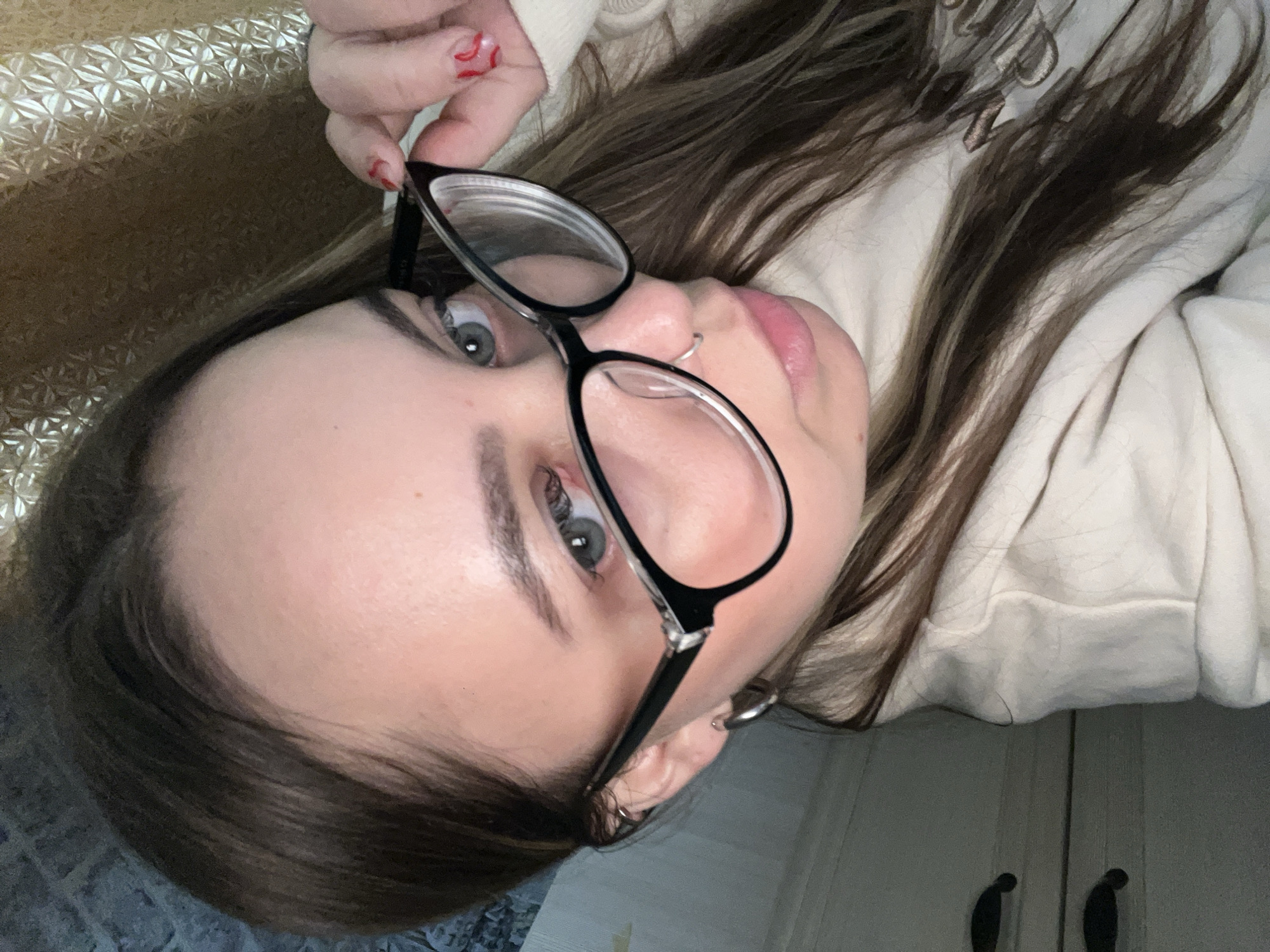 A close-up, angled portrait features a young person, likely female, indoors. The subject has medium-brown hair and is looking directly at the camera, holding black-framed glasses with her right hand near her temple. Her left eye is visible through the lens, showing a light-colored iris, complemented by defined eyebrows and mascara. A small silver hoop piercing adorns her right nostril, and her lips are slightly parted. Her right hand, visible in the upper left, has red nail polish, which shows some wear. She is dressed in a light-colored, possibly beige or off-white, long-sleeved top, likely a hoodie or sweatshirt.
The background consists of two main elements. To the left, behind her head, is a golden or beige textured surface with a repeating, abstract pattern. To the lower right, part of a light-colored wooden furniture piece, possibly a cabinet or dresser, is visible, featuring two dark, curved handles. The lighting suggests an indoor setting, without strong natural light. The subject's expression is neutral and direct, implying a casual moment. No text is visible in the image. The scene evokes a relaxed atmosphere, potentially within a home in Novopokrovskaya, Russia.