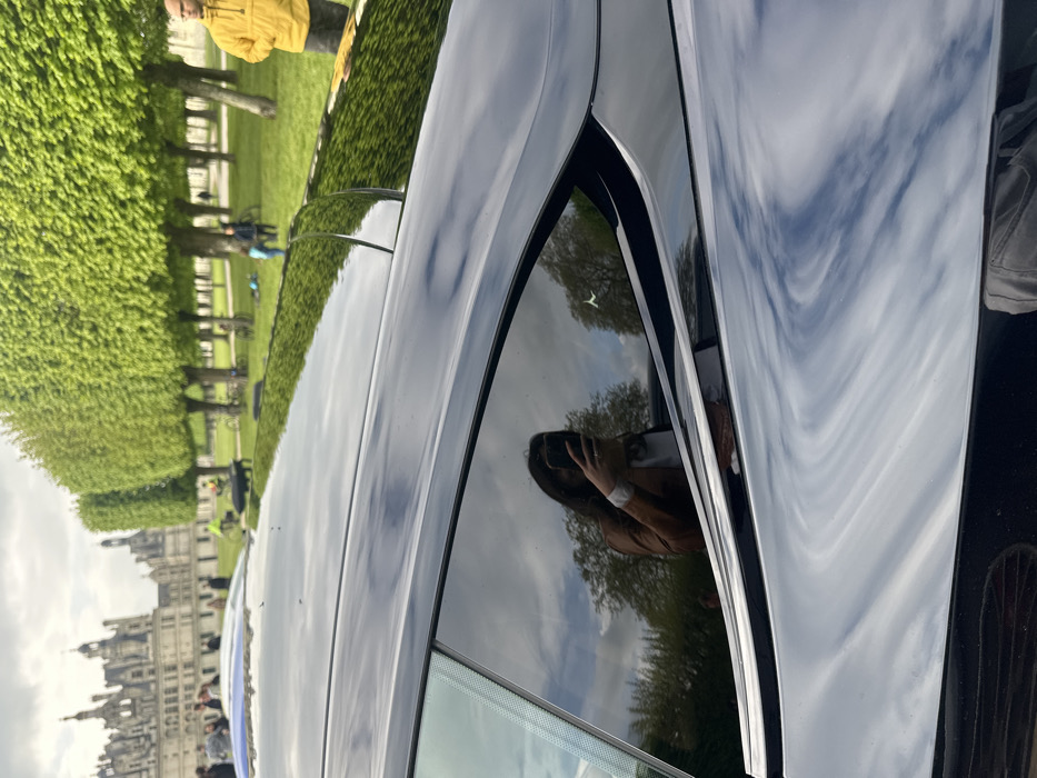 The image shows the rear side of a black car parked in front of the Chateau de Chambord, a large, historic castle in Chambord, France. The car is facing the castle, which is visible in the background. The car's windows are tinted, and the reflection of a woman taking a selfie with a phone is visible in the rear window. The woman is wearing a white shirt and a brown jacket. The car is parked on a grassy field, with a line of trees behind the car. The trees are all green and leafy, and the sky is cloudy. There are a few people in the background, including one man in a yellow jacket. The coordinates of the Chateau de Chambord are approximately 47.616°N, 1.519°E. The image is shot at a slight upward angle. The car is in focus, with a soft focus on the background. The image is composed in a way that emphasizes the car and its reflection. The image is shot in natural light.