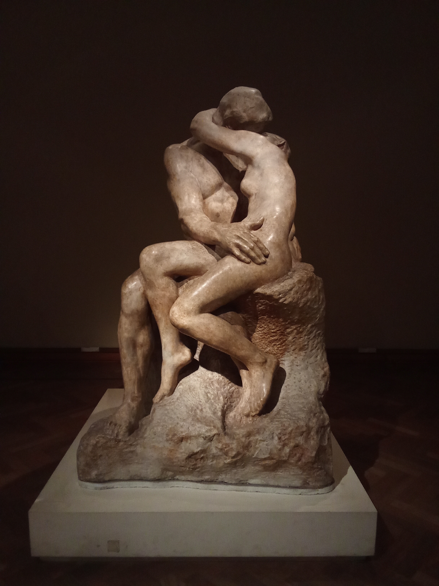 The image shows a sculpture of two embracing figures, likely "The Kiss" by Auguste Rodin. The sculpture is made of a light-colored material, possibly marble or bronze, and depicts a man and a woman in an intimate embrace. They are seated on a rough, rocky base. The figures are rendered in a realistic style, with smooth, flowing lines. The sculpture is displayed on a white rectangular pedestal in a dimly lit room, suggesting it is in a museum or gallery. The background is dark and out of focus.
Based on the provided context, this sculpture is located in the Museo Nacional de Bellas Artes in Buenos Aires, Argentina.