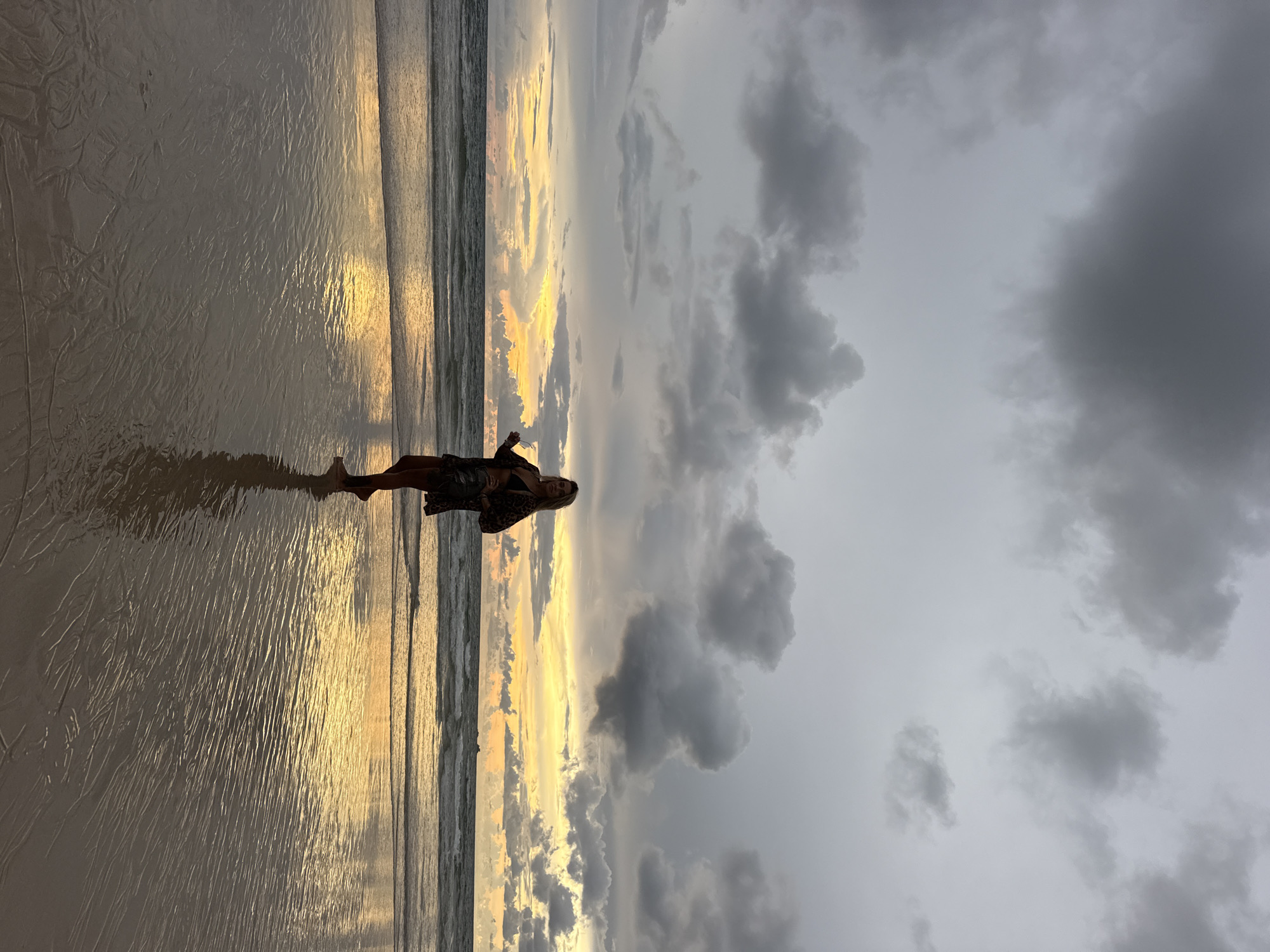 A full-body, vertical shot captures a woman standing on a wet beach during twilight. The woman, with blonde hair and wearing a dark patterned outfit and sunglasses, is positioned in the center of the frame, facing left and looking towards the ocean. Her reflection is visible on the wet sand. The beach is smooth with patterns from receding waves. The ocean stretches to the horizon, where a band of warm yellow and orange light, characteristic of a sunset or sunrise, is visible through dark, dramatic clouds. The sky is filled with heavy, grey clouds, with some breaks allowing the colorful light to peek through. The overall scene is one of natural beauty and tranquility. The location is the beach in Choeng Thale, Thailand.