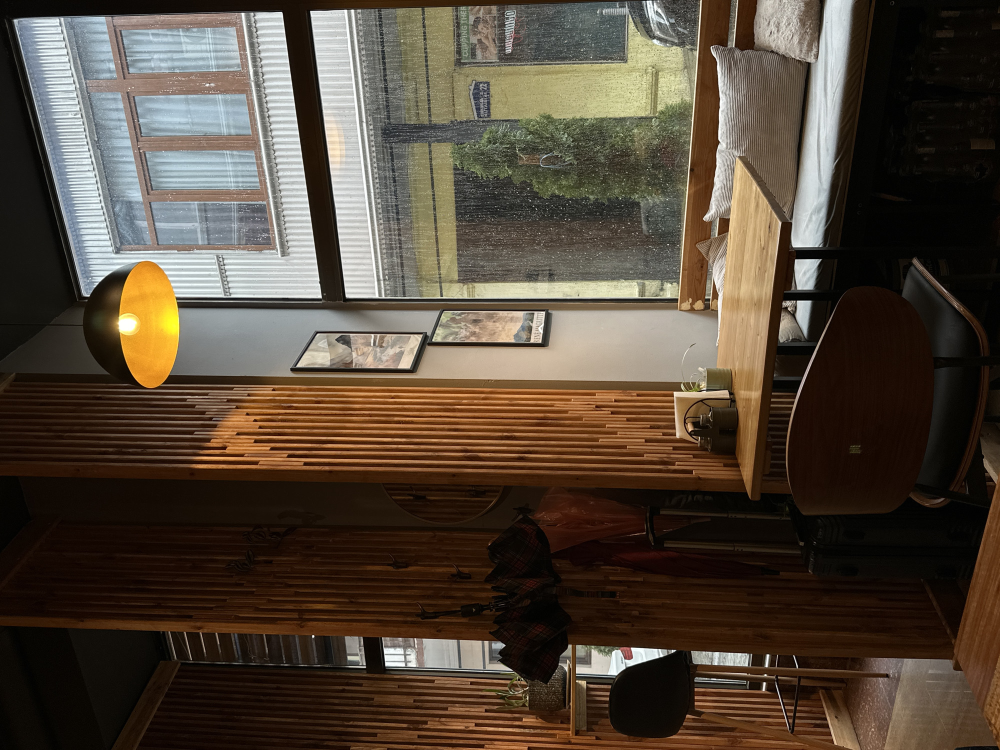 The image shows the interior of a cafe or restaurant in Batumi, Georgia. Rain is visible on the window, obscuring the view of buildings across the street. Two framed pictures hang on the wall, and a gold-colored pendant lamp illuminates a section of the room. The wall and ceiling are covered with horizontal wooden slats. An umbrella hangs on the wall. A table and chairs are arranged for customers, and a cushioned bench is positioned near the window.