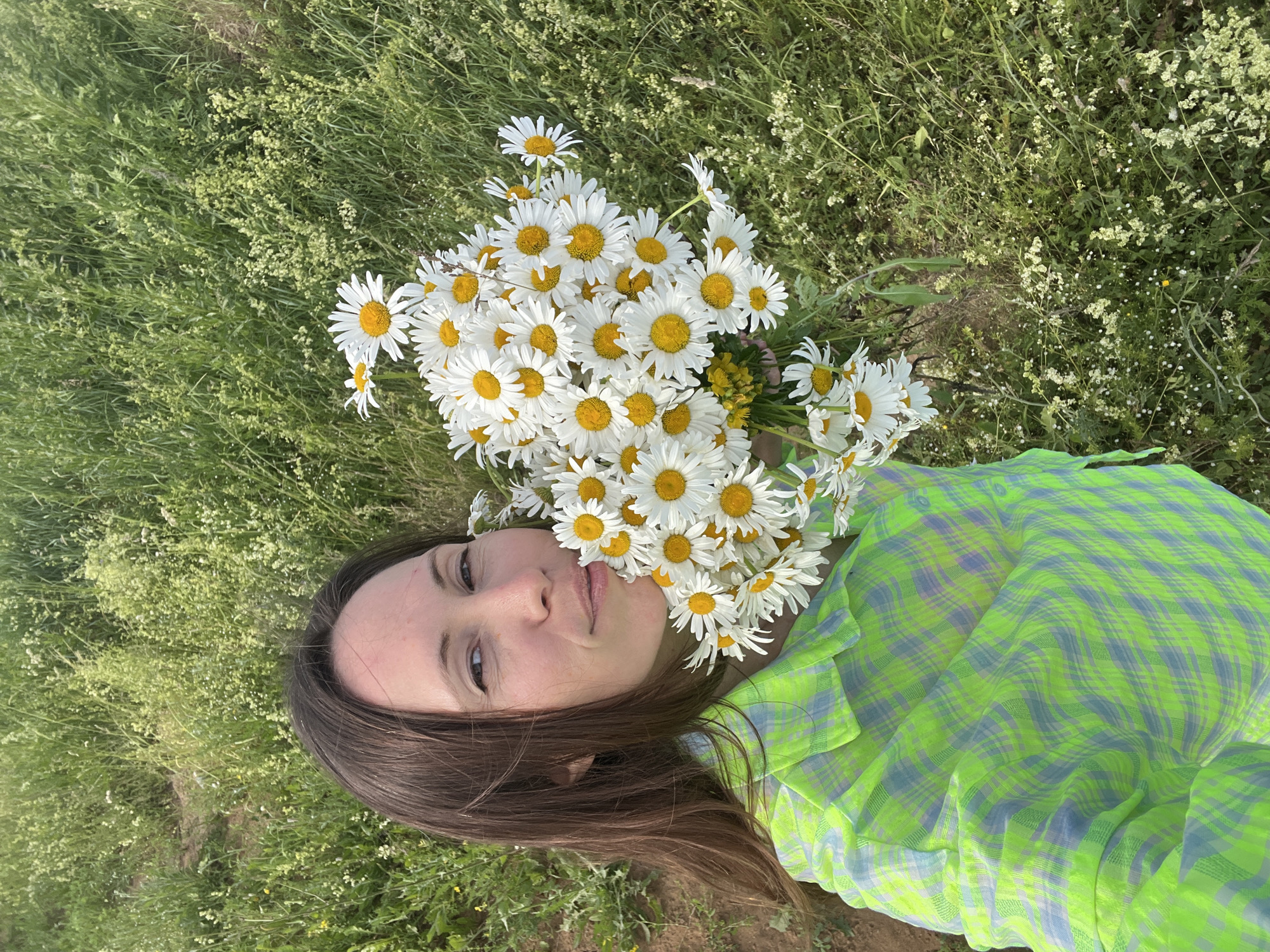 A woman with long, dark brown hair and fair skin is holding a large bouquet of white daisies with yellow centers. She is wearing a green and blue plaid shirt. She is looking at the camera with a slight smile. The background is a field of green grass and wildflowers. The scene is outdoors. The location is described as Пигозино, Russia.