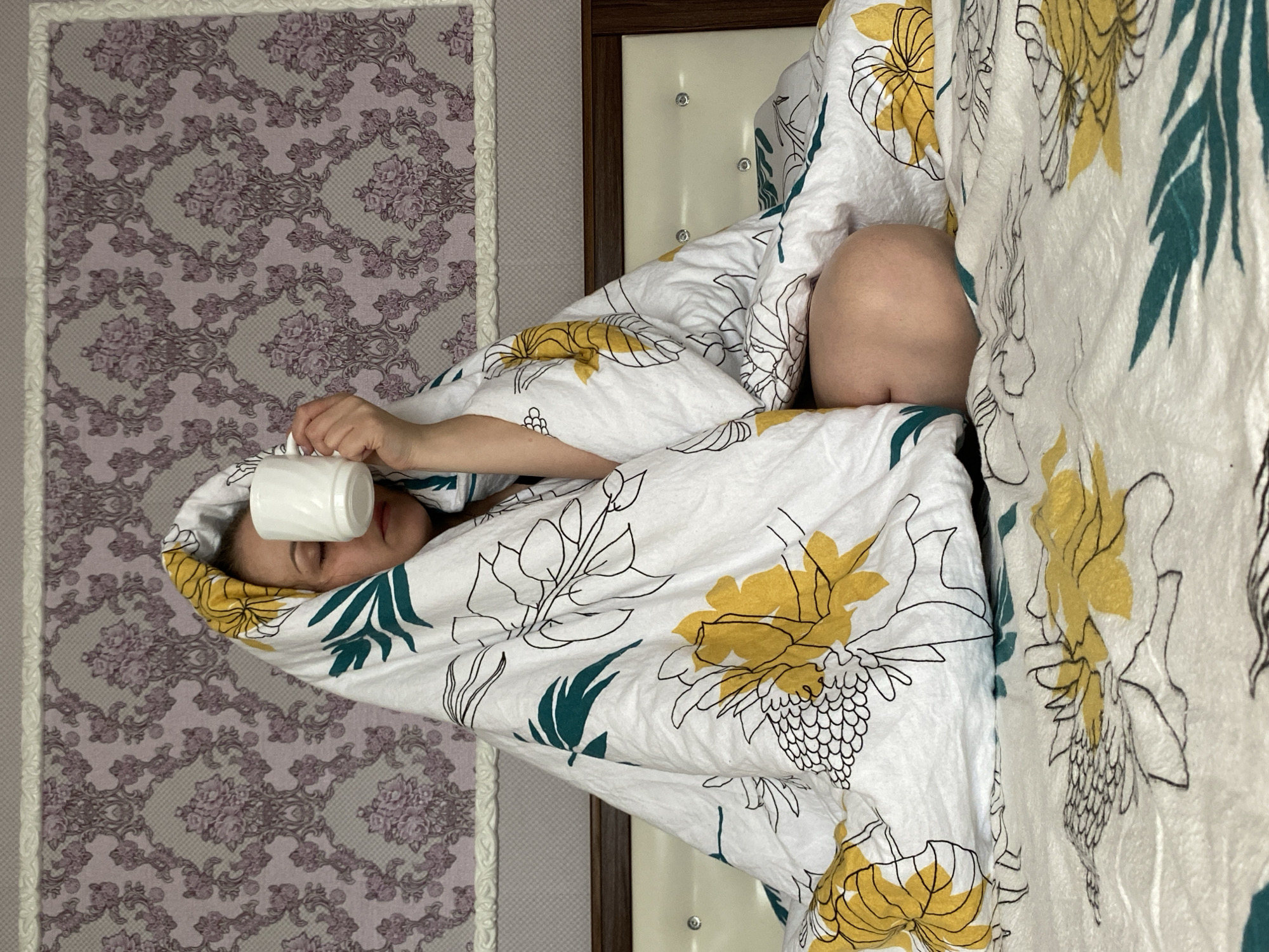 The media file presents an intimate indoor scene, possibly a bedroom, captured from a top-down perspective and rotated 90 degrees counter-clockwise.
A person, likely a child or young adult, is lying in bed, mostly enveloped by a white duvet. Their head is visible, with eyes closed, suggesting rest or sleep. One hand is holding a plain white mug, which is gently placed against their forehead or eye area. The duvet features a distinctive pattern of yellow and black-outlined floral and leaf designs, complemented by teal-colored accents.
Behind the person's head, a section of a bed headboard is visible, characterized by a dark wooden frame and a padded white insert with decorative buttons. To the left of the bed, the wall is adorned with elegant purple and grey floral damask wallpaper, separated from an unpatterned wall section by a textured white decorative border.
The activity depicted is one of quiet repose, with the person appearing to be sleeping or resting, possibly due to feeling unwell, indicated by the closed eyes and the mug's placement. The setting clearly indicates a private residence in Markovskii, Russia. The time of day and external weather conditions cannot be determined from this indoor view. There is no visible text in the image.
