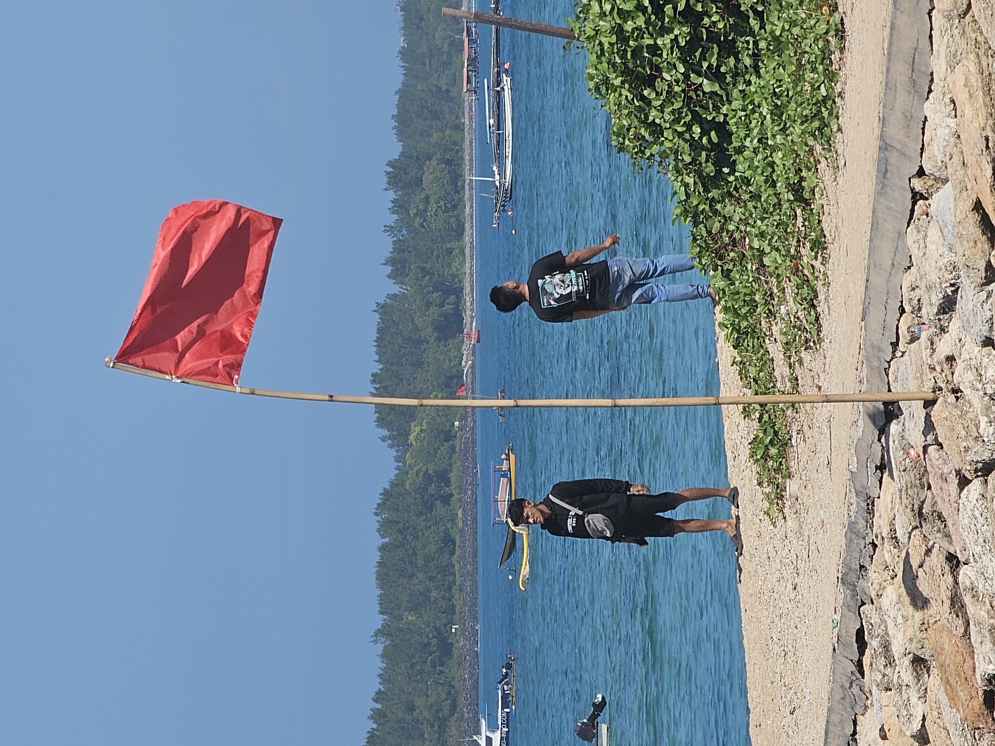 The image shows a coastal scene in Denpasar, Indonesia, with a red flag flying prominently on a bamboo pole in the foreground. Two men are standing on a sandy embankment overlooking the ocean. One man, on the left, is wearing a black hoodie, black shorts, and a crossbody bag. The other man, on the right, is wearing a black t-shirt and blue jeans. The ocean is calm and blue, with several boats visible in the distance. A lush green bush is growing on the right side of the embankment. The background features a tree line and a clear blue sky. The overall scene suggests a sunny day at the beach.