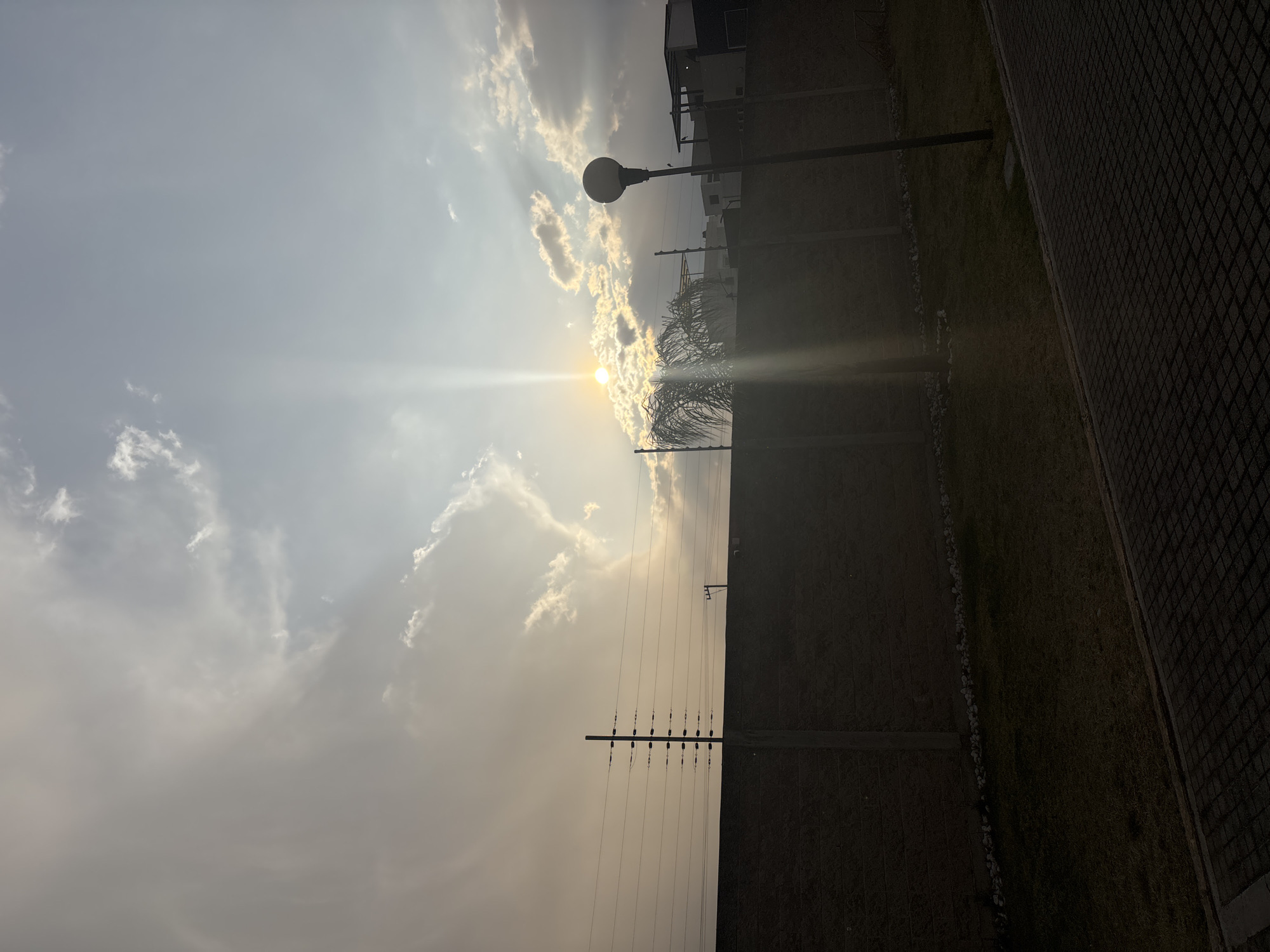 The image depicts a view towards the sky, with the sun obscured by clouds. In the foreground, there is a wall with electrical wires and a lamppost. To the right, there is a paved area. The location is not Curitiba, Mexico, as Curitiba is a city in Brazil.