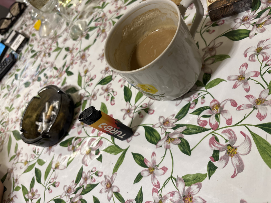 A white mug filled with coffee sits on a table with a floral tablecloth. An ashtray filled with cigarette butts and a black and orange "Turbo" lighter are nearby. A box of what appears to be Chesterfield cigarettes is partially visible in the upper right corner. Glasses and other objects are out of focus in the upper left corner, completing the scene of a casual, lived-in space.